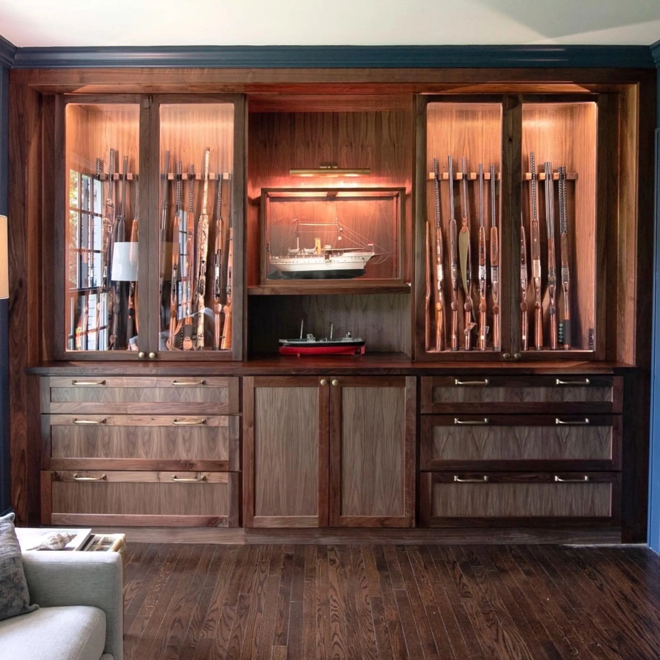 A large wooden cabinet with glass doors displaying antique rifles and a framed model ship on top. There is a small model boat on the shelf below the framed ship. The cabinet has multiple drawers and doors, and the room has a hardwood floor.