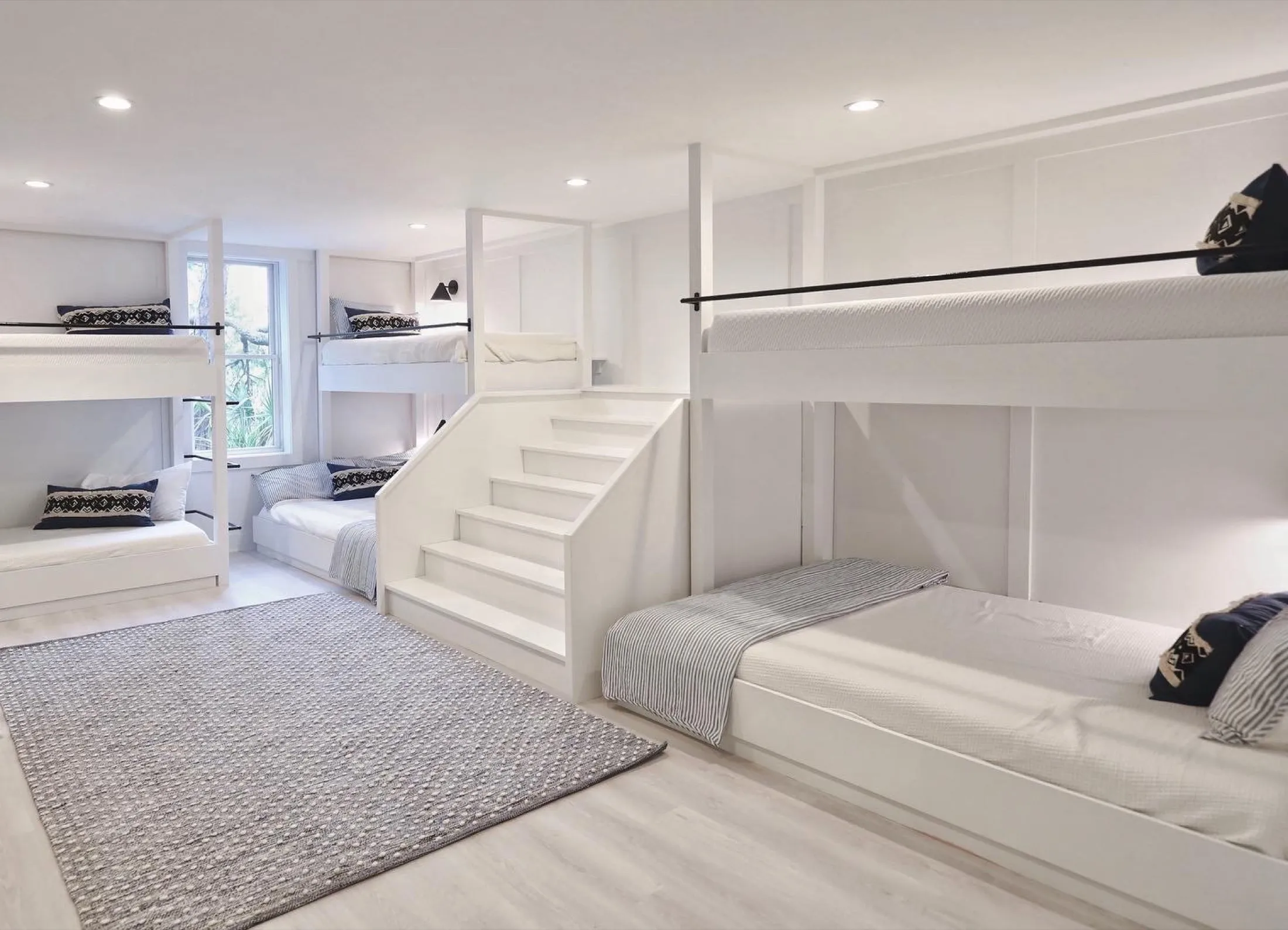 Room with white walls and ceiling, featuring multiple bunk beds with black and white pillows and bedding, a window with trees outside, a patterned rug, and white flooring.