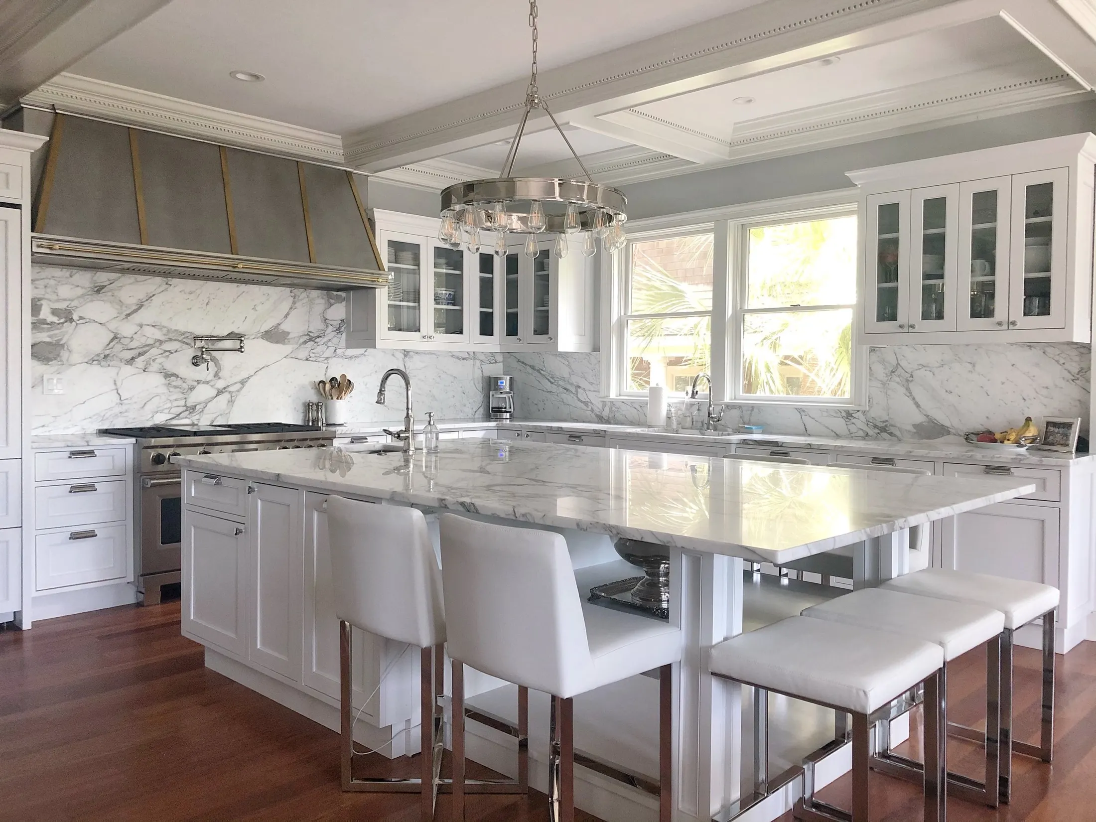 Modern kitchen with white cabinets, marble countertops, a large marble island, stainless steel appliances, glass-front cabinets, and hardwood floors. There are three white barstools at the island and a chandelier hanging overhead.