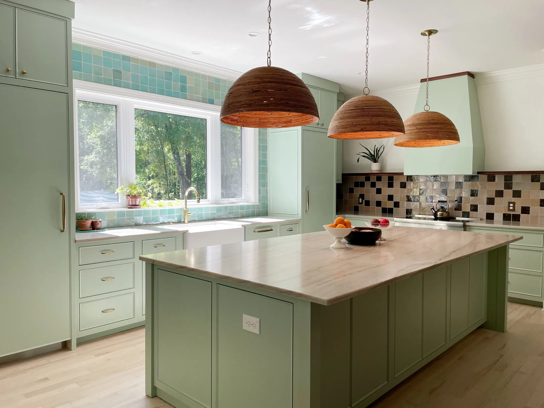 A bright kitchen with green cabinets, a large island with a beige marble countertop, three hanging wooden pendant lights, and a large window overlooking greenery. The backsplash is a mix of square tiles in neutral tones.