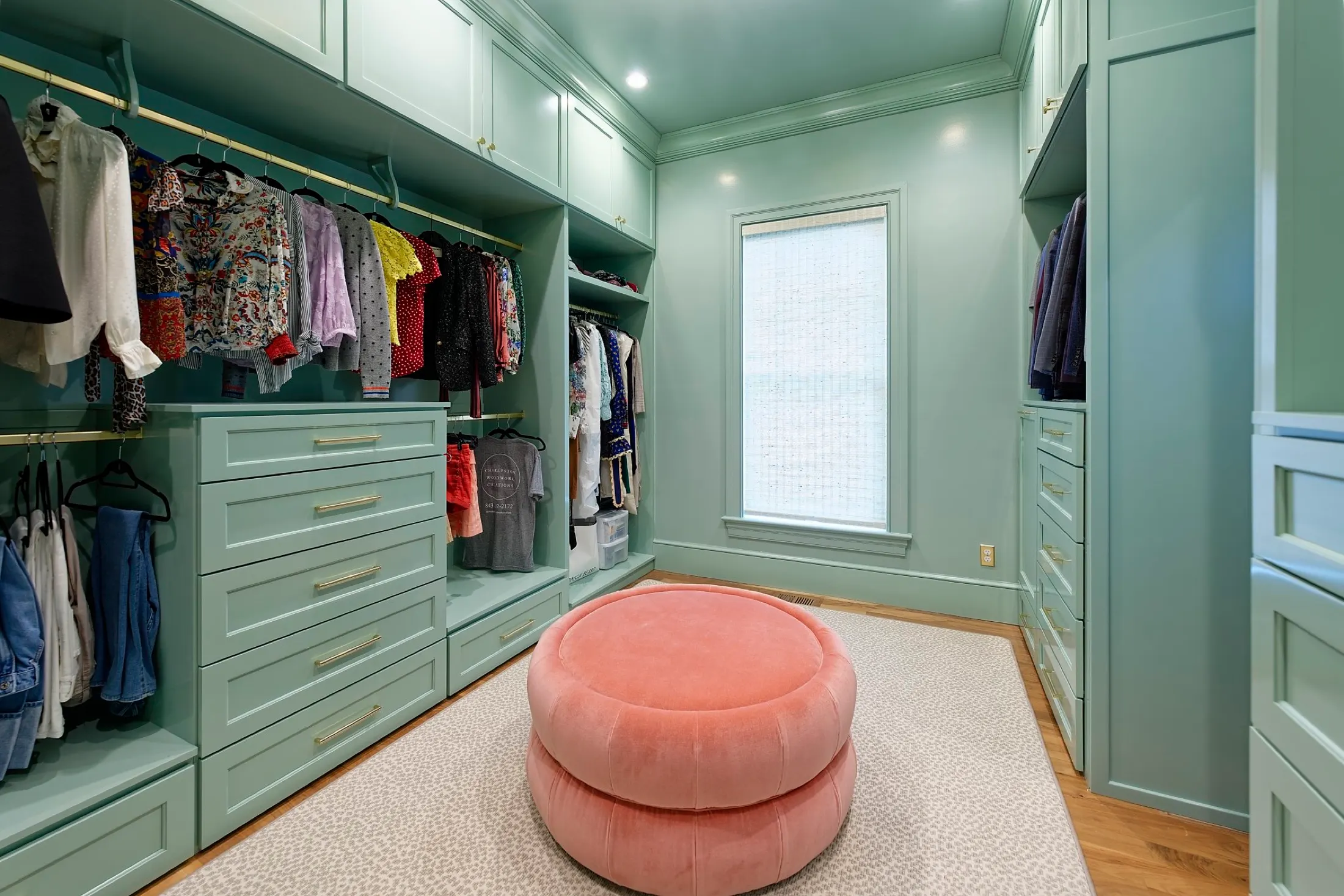 A walk-in closet with green built-in cabinets and drawers, hanging clothes, a pink ottoman in the center, a window with a white shade, and wooden flooring.
