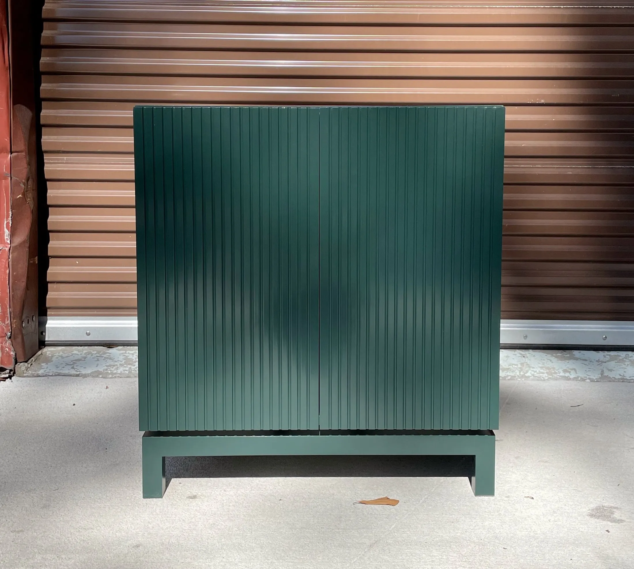 Front view of a green wooden cabinet with vertical slats, placed outdoors on a concrete ground with a brown metal garage door in the background.