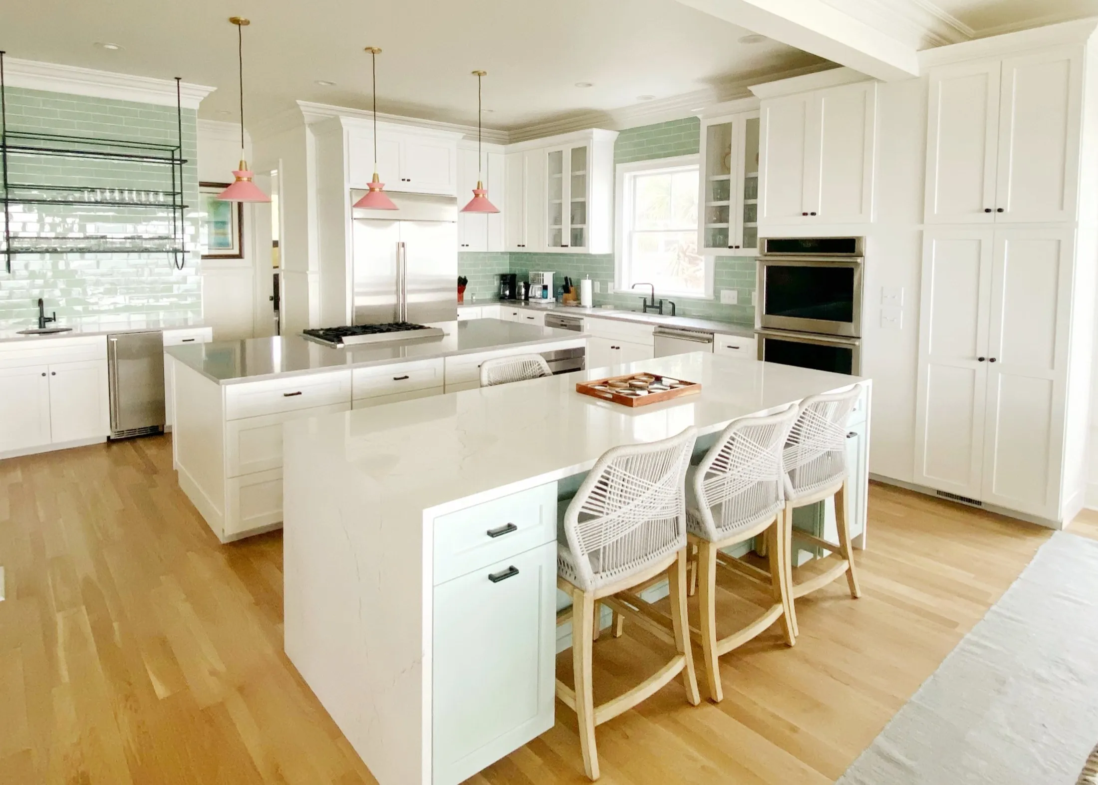 Modern white kitchen with a large central island, three white chairs, wood flooring, hanging pendant lights, and green tiled backsplash.