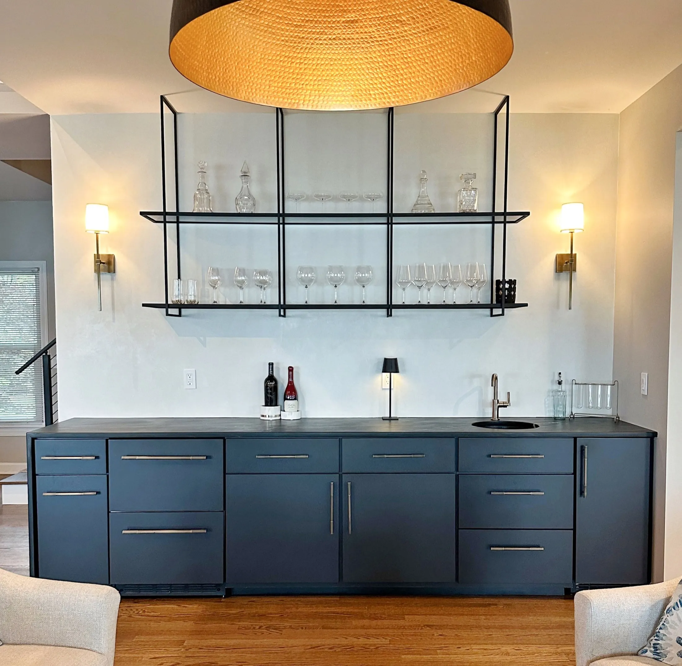 Modern bar area with blue cabinetry, black countertop, glassware on black metal shelves, small black table lamp, and wall-mounted lights.