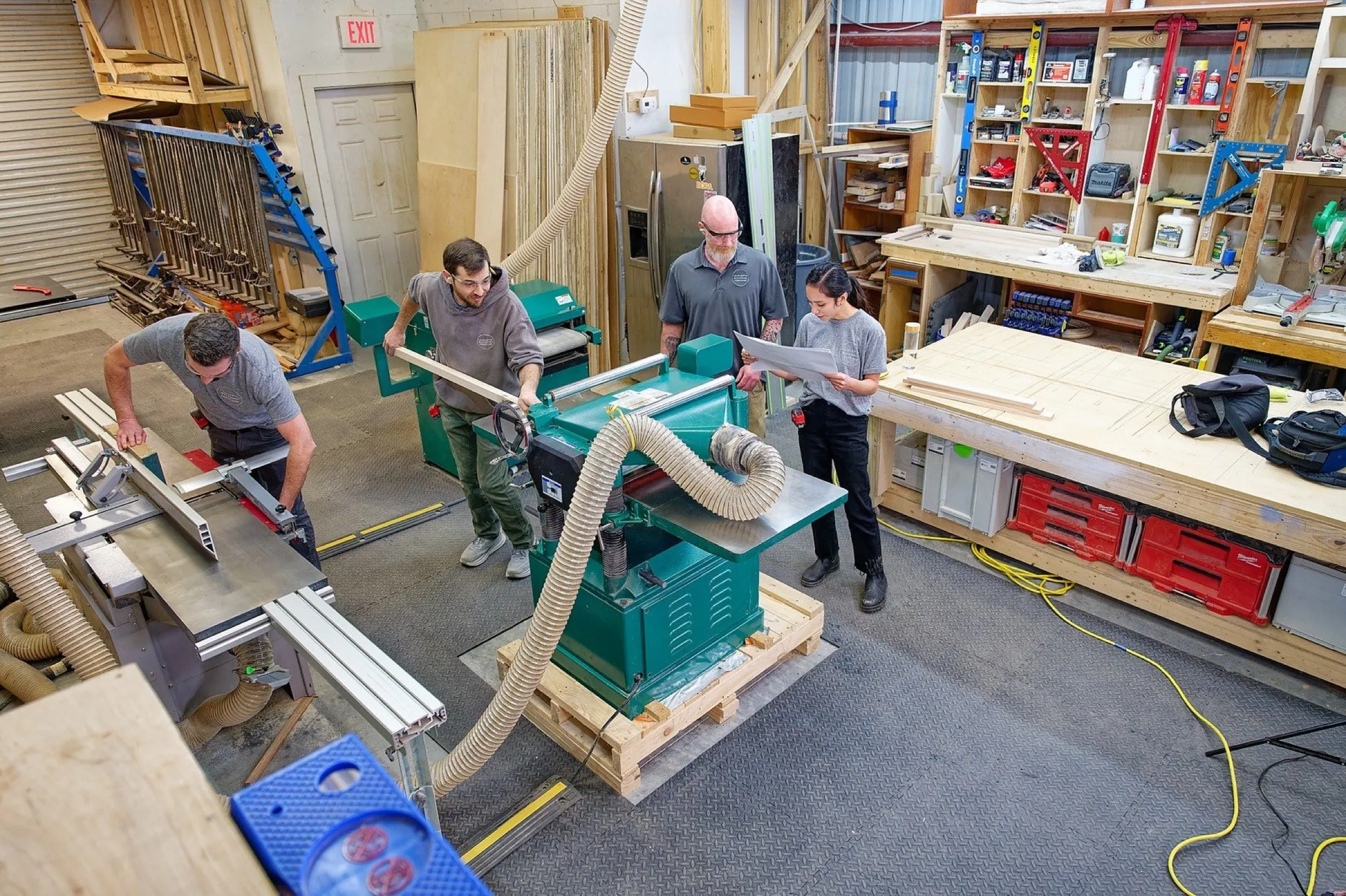 Four people working in a woodworking shop. One person is using a table saw, two are operating a large green machine, and one is reading a blueprint. The shop has various woodworking tools, lumber, and storage shelves along the walls.