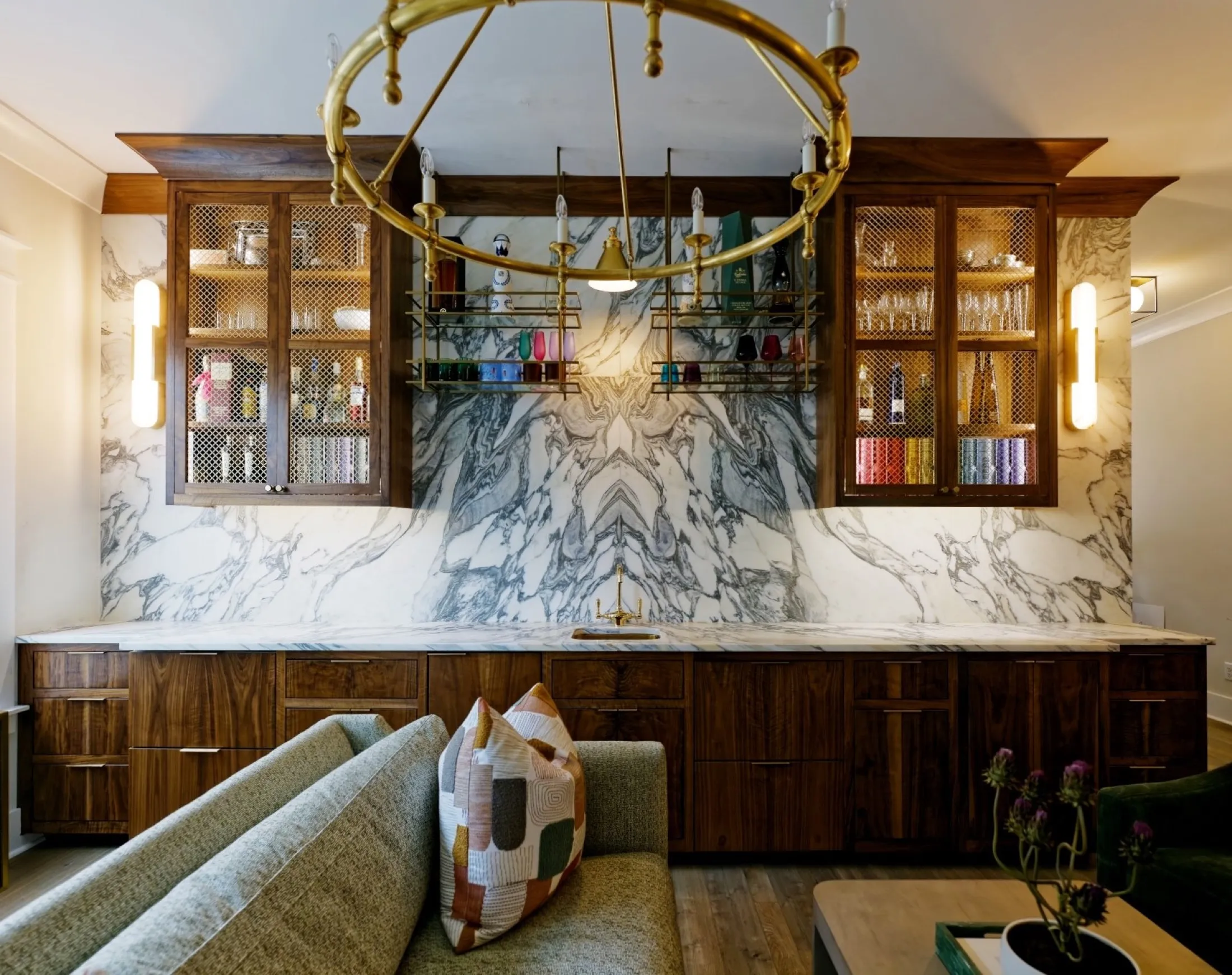 A home bar with walnut cabinetry, glass-front uppers, a dramatic book-matched marble backsplash and a brass chandelier.