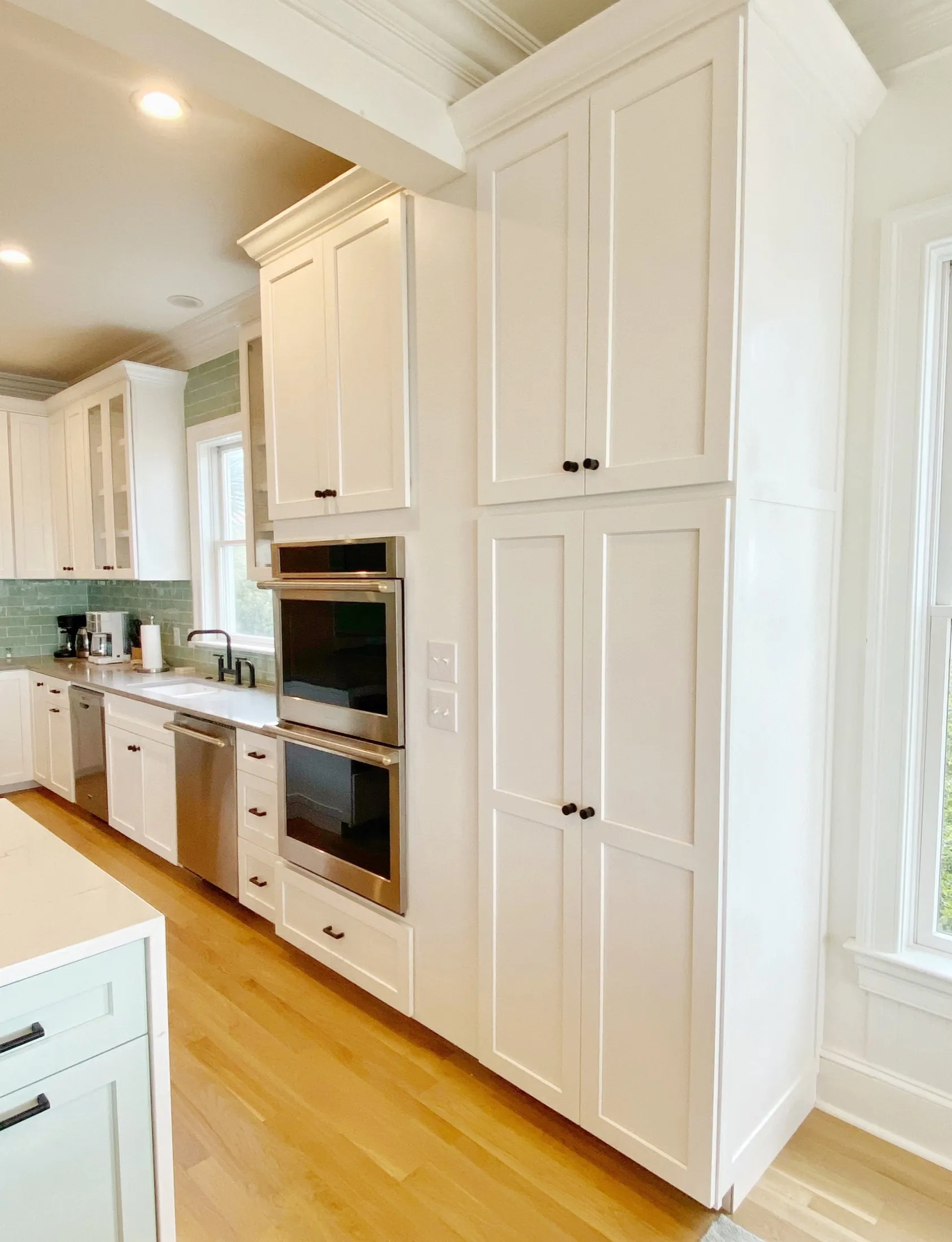 A modern kitchen with white cabinets, wood flooring, a double oven, a sink under a window, and green tiled backsplash.