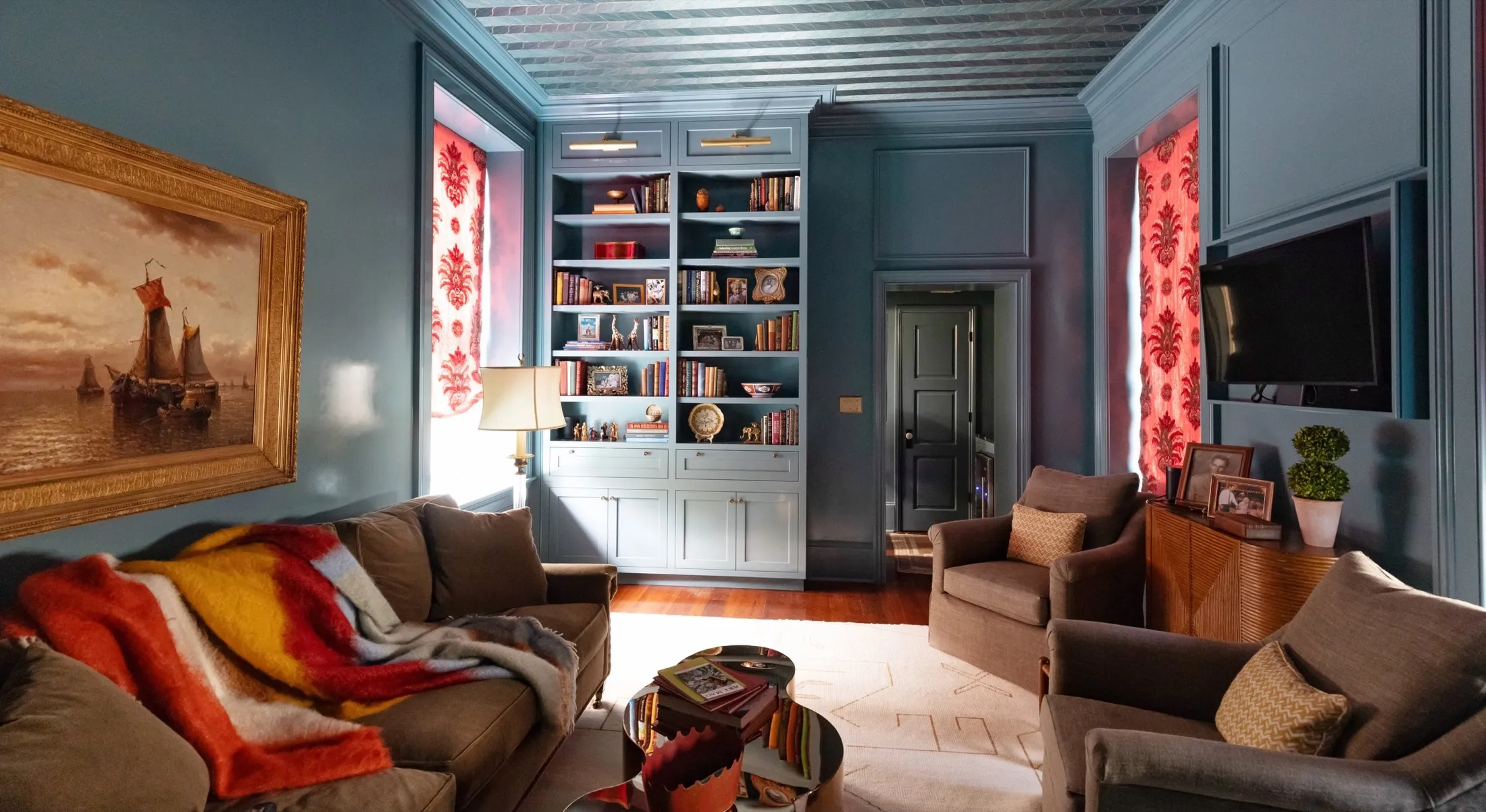 Living room with blue walls, a large bookshelf, a painting of sailboats, a brown sofa with a colorful blanket, two armchairs with pillows, a wooden sideboard with photos and a plant, a wall-mounted TV, red patterned curtains, and a white rug.