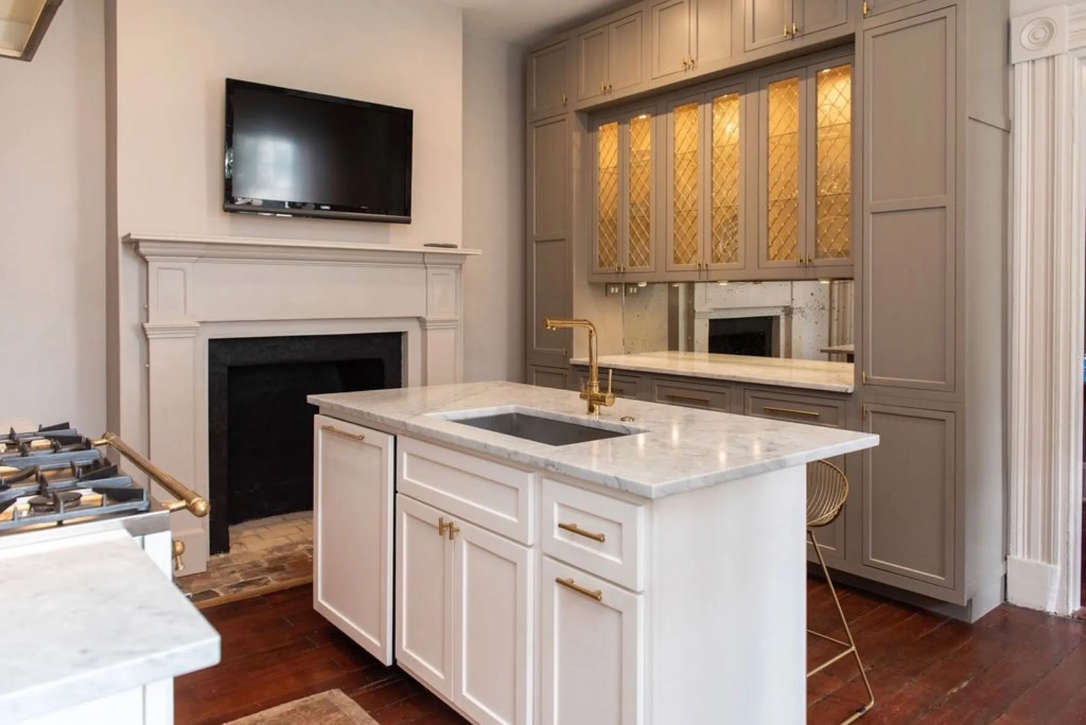 Modern kitchen with white marble countertops, gold fixtures, gray cabinets, and a flat-screen TV above a fireplace.