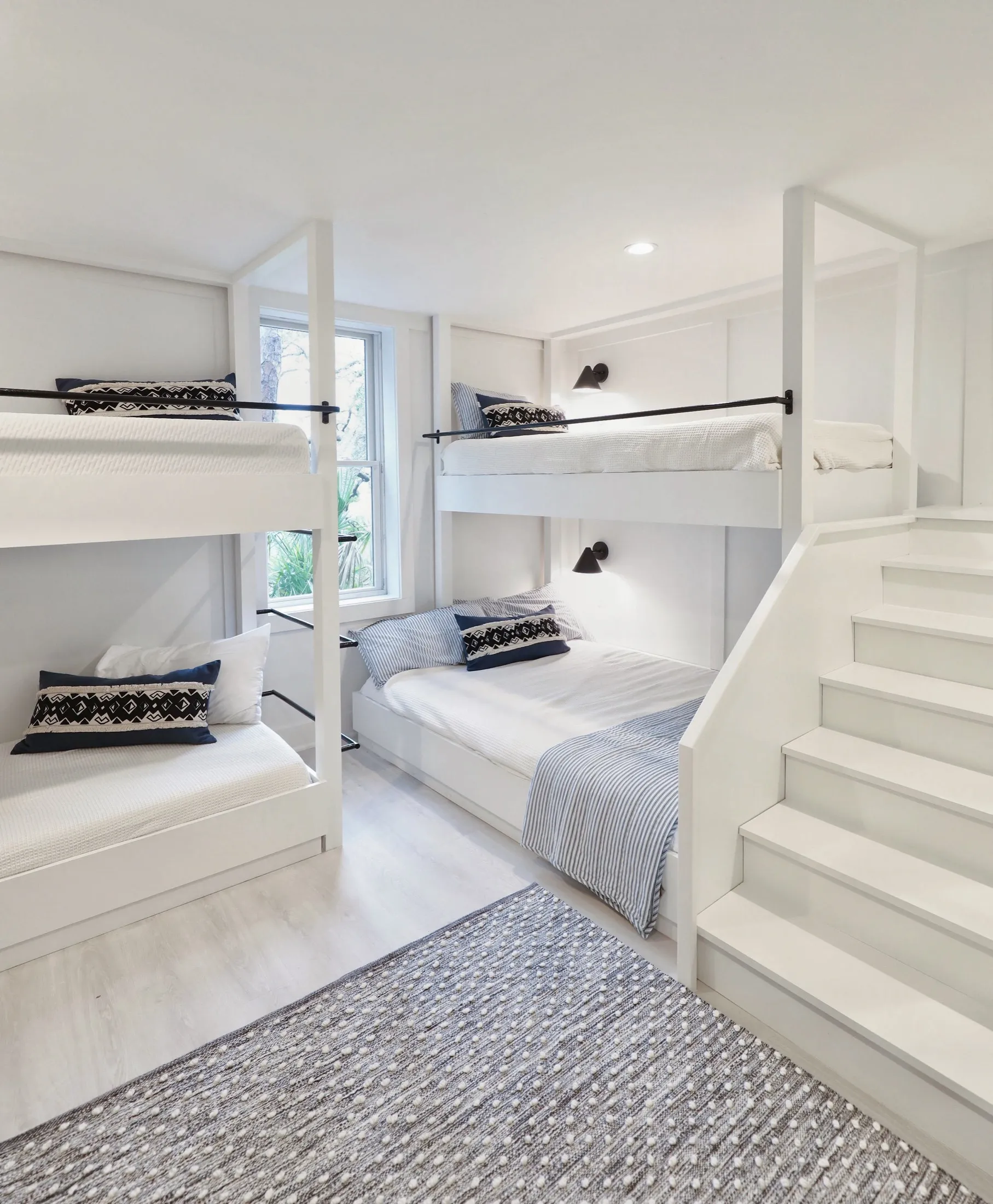 Cozy bedroom with white bunk beds and a double bed, black wall-mounted lamps, and a window with greenery outside, decorated in neutral tones.
