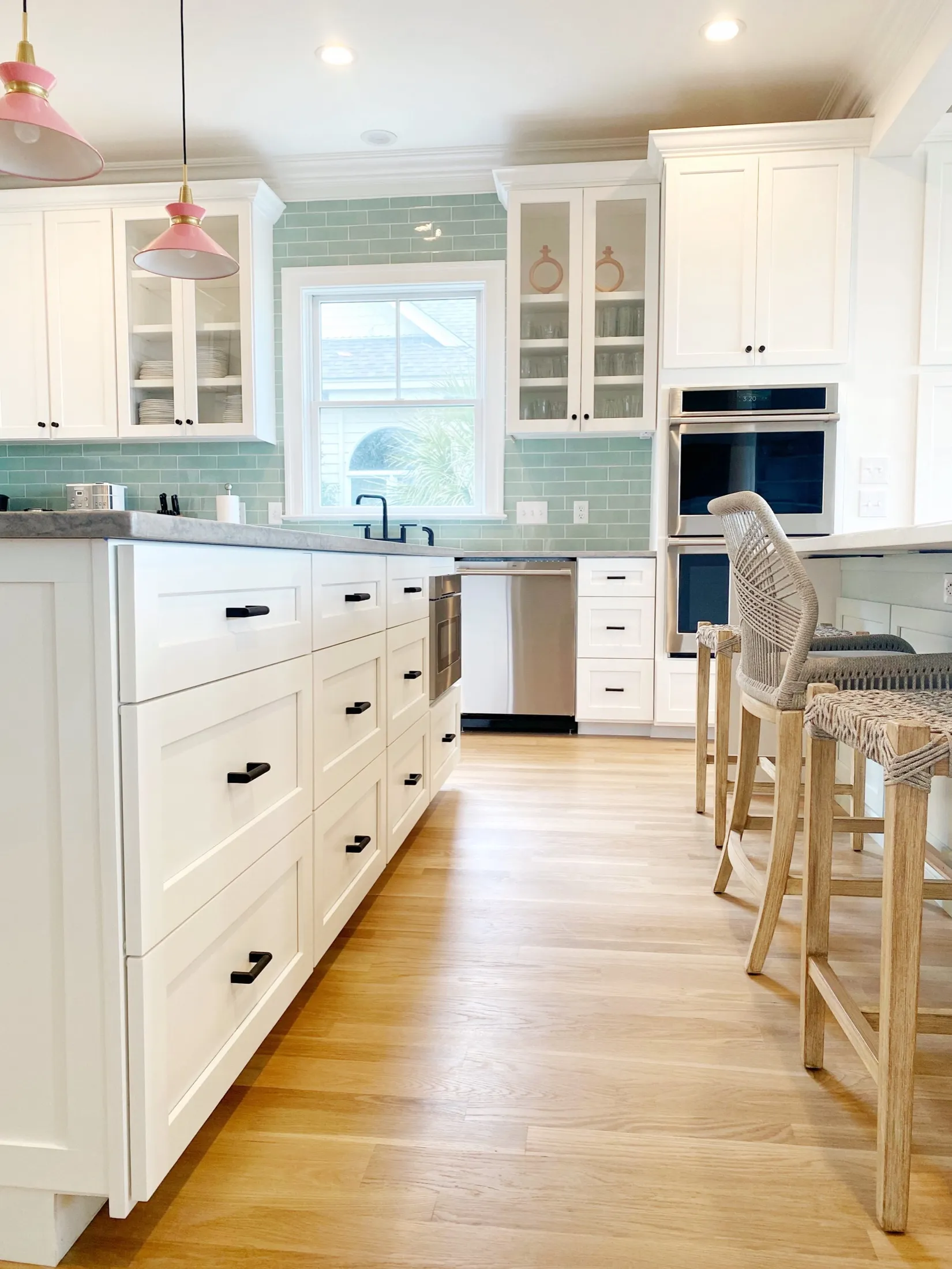 Bright, modern kitchen with white cabinets, wooden flooring, and a light green tiled backsplash. Includes a window with a view outside, a stainless-steel dishwasher, and built-in oven. There are pink pendant lights hanging above the counter and a gra