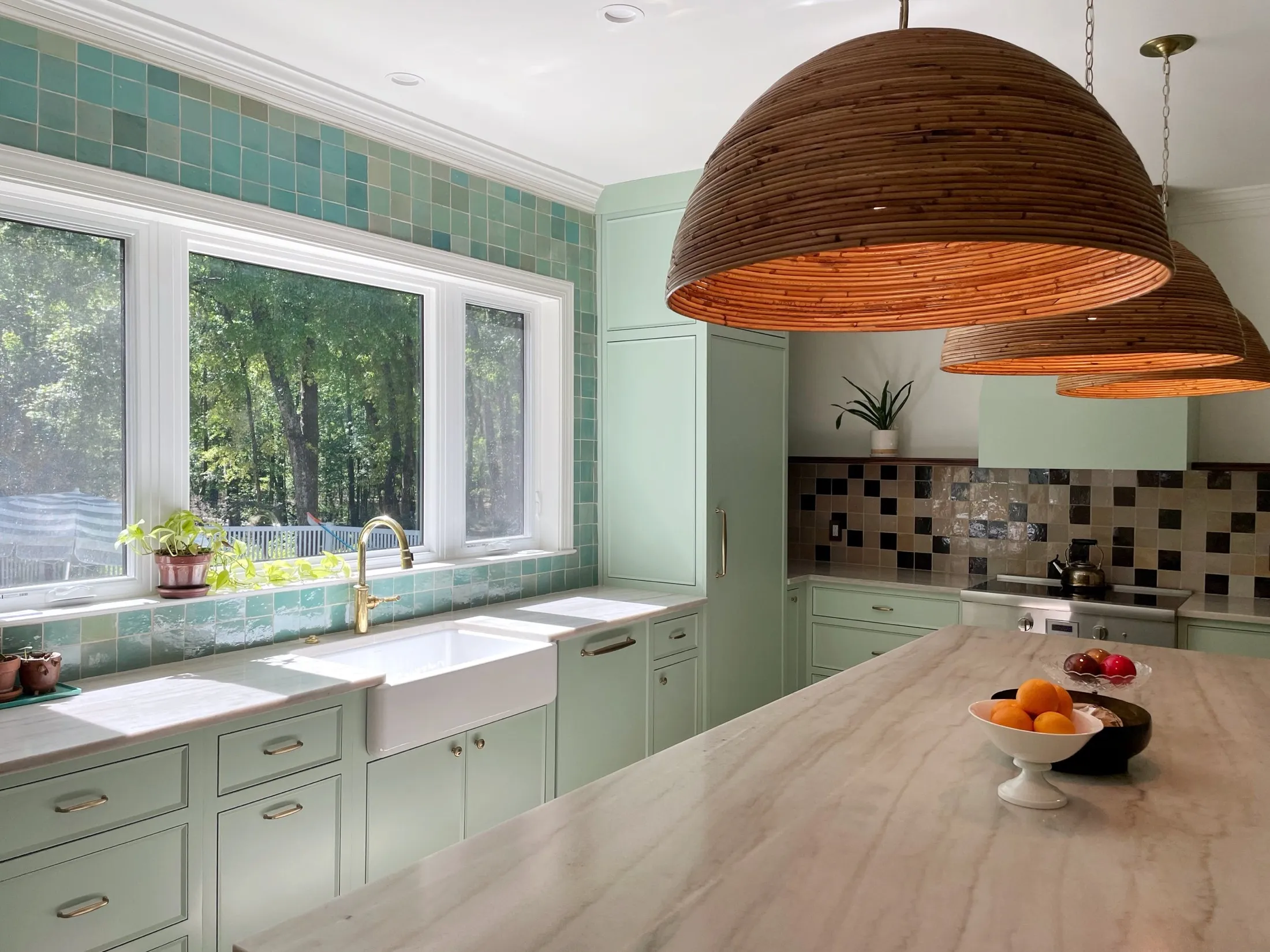 Bright kitchen with light green cabinets, a white farmhouse sink, large window with greenery outside, pendant wooden light fixtures, tiled backsplash, and a marble countertop with fruit bowls.