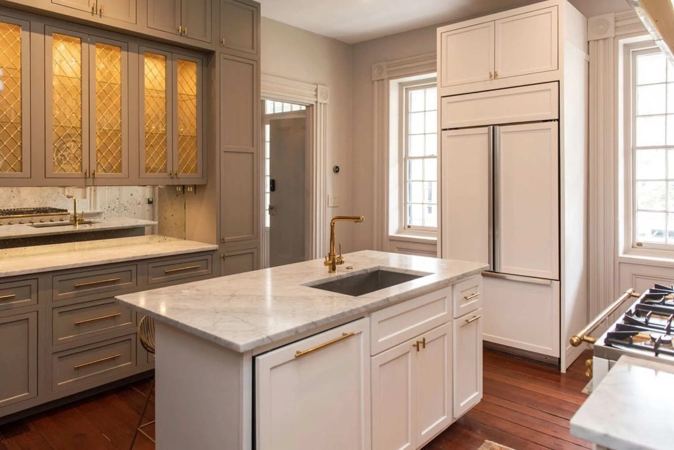 A modern kitchen with white and gray cabinets, marble countertops, a gold faucet, a built-in refrigerator, and large windows.