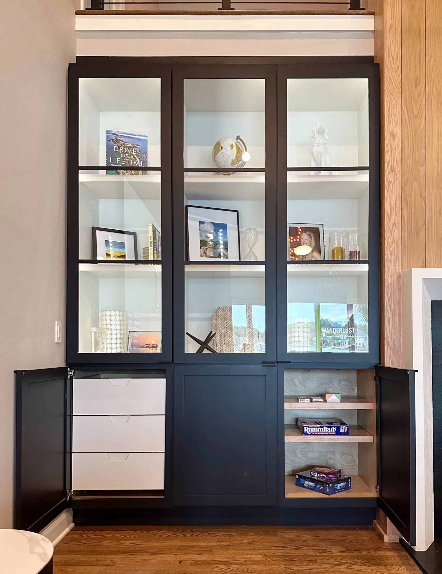 A built-in cabinet with glass doors containing books, framed photographs, and decorative items, with closed black doors on the bottom left and open shelves on the bottom right showing board games and boxes.