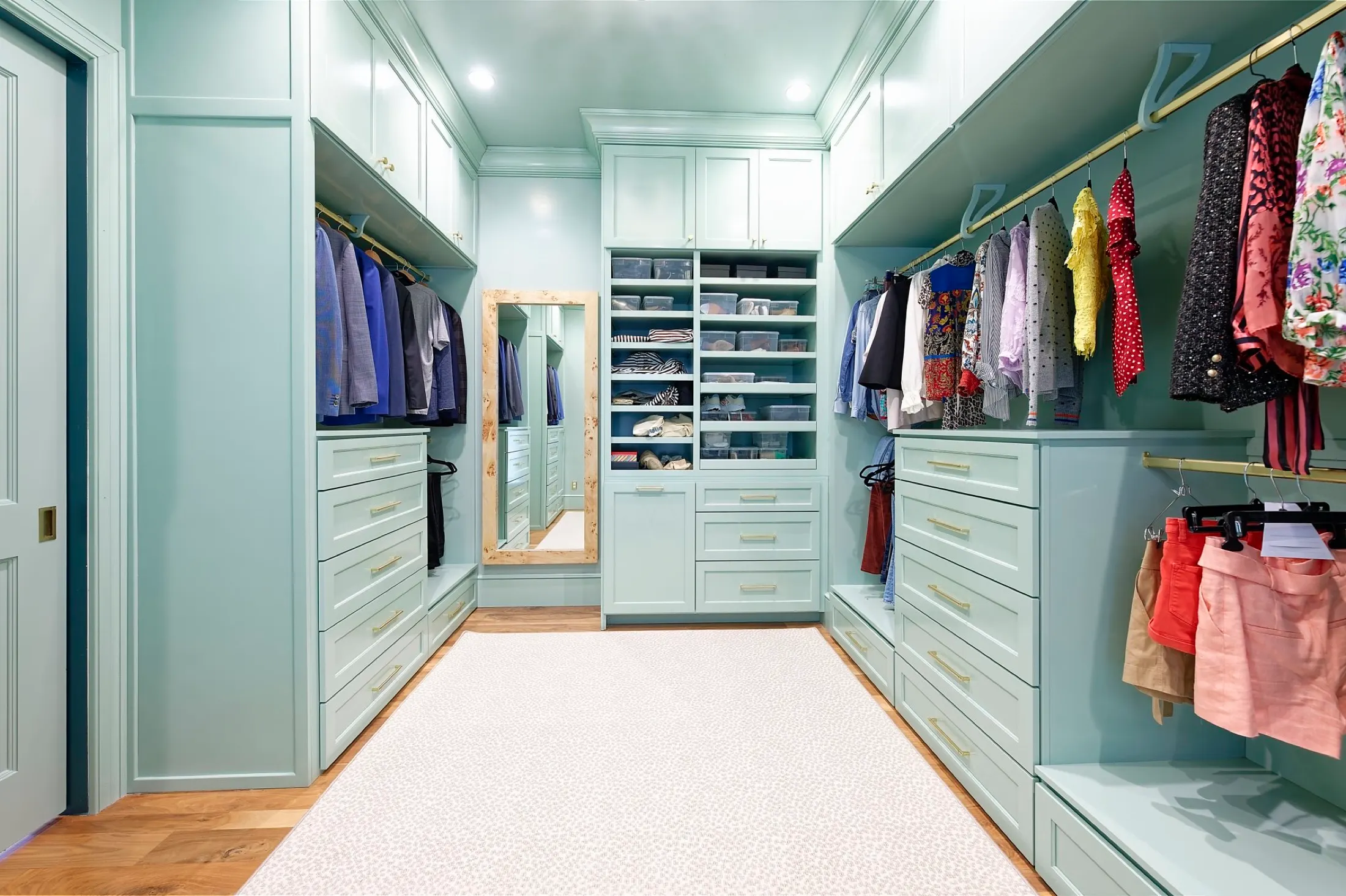 A spacious walk-in closet with mint green cabinetry and gold handles, featuring clothing racks with various garments, a full-length mirror, and organized storage drawers and shelves for shoes and accessories.