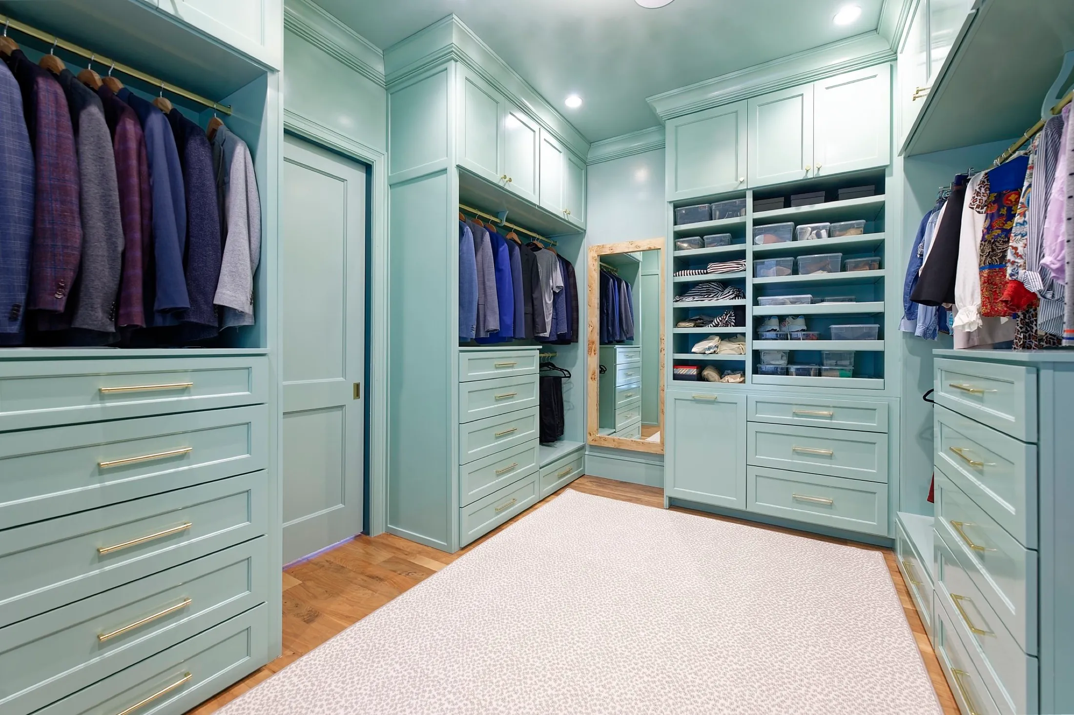A spacious walk-in closet with light teal cabinetry, gold handles, and wooden flooring. The closet features hanging clothes, drawers, shelves with folded items, and a full-length mirror.