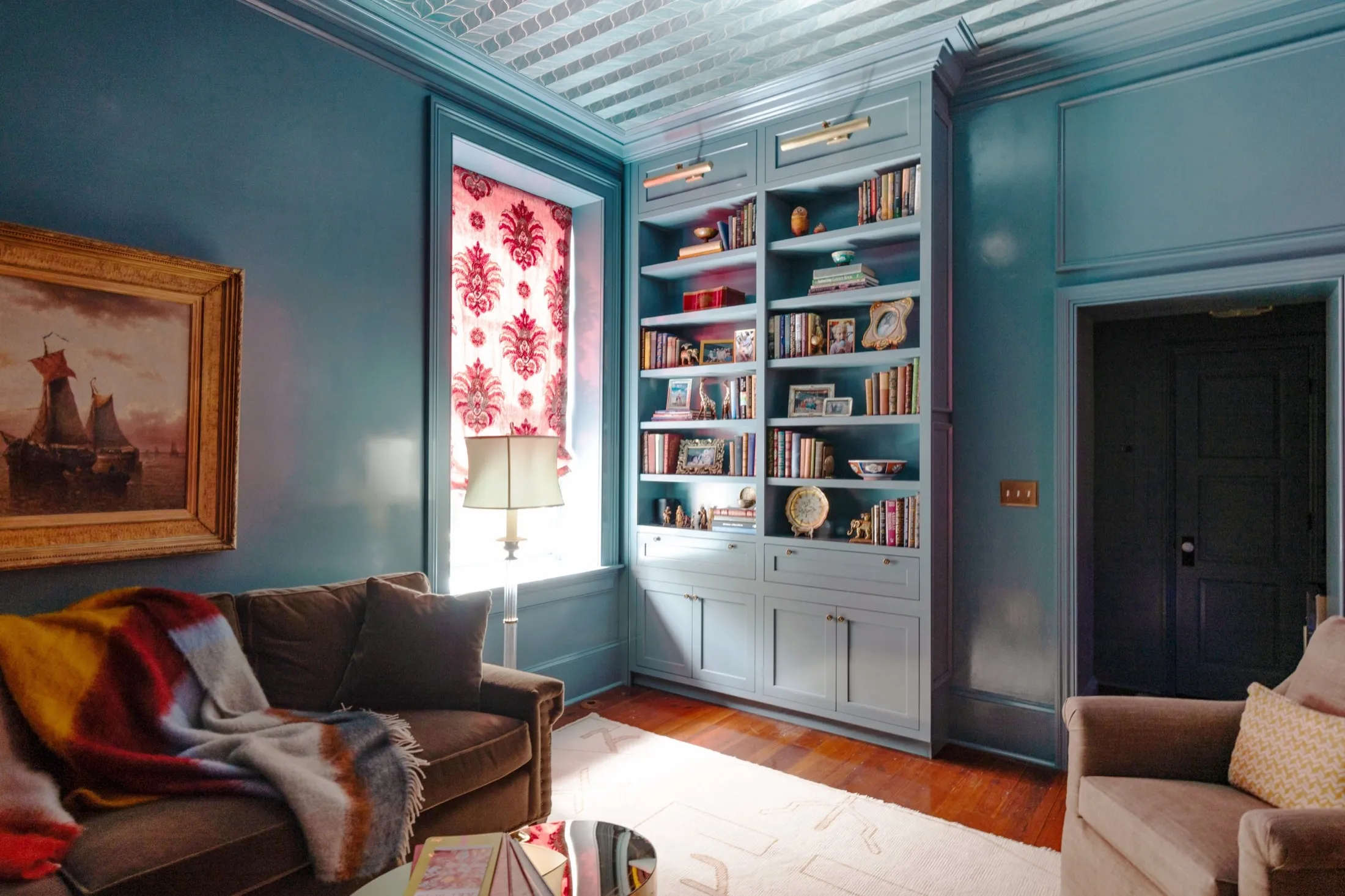 Living room with blue walls, large built-in bookshelf filled with books and decorative items, window with patterned red and pink curtains, beige armchair, dark brown sofa with colorful blanket, floor lamp, painting of ships, and wooden flooring.