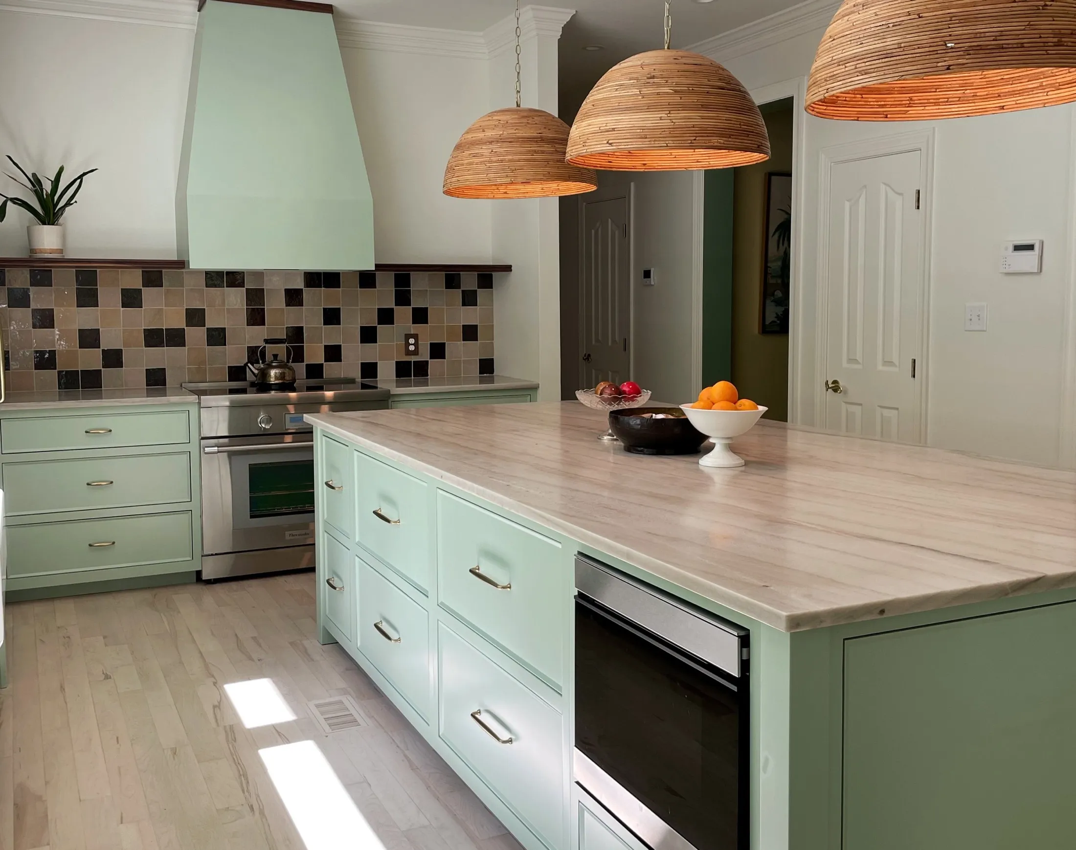 A kitchen with mint green cabinets, a marble island, a stainless steel stove, a patterned tile backsplash, three hanging wooden pendant lights, a bowl of fruit on the island, and a potted plant on the counter.