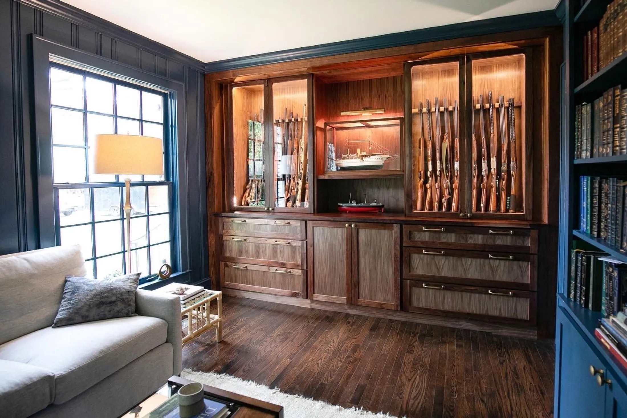 A living room with dark wooden walls and flooring, a large window with a beige lampshade lamp next to a gray sofa with a dark gray pillow. There is a light blue bookshelf on the right side and a wooden display cabinet with collection of rifles and mo