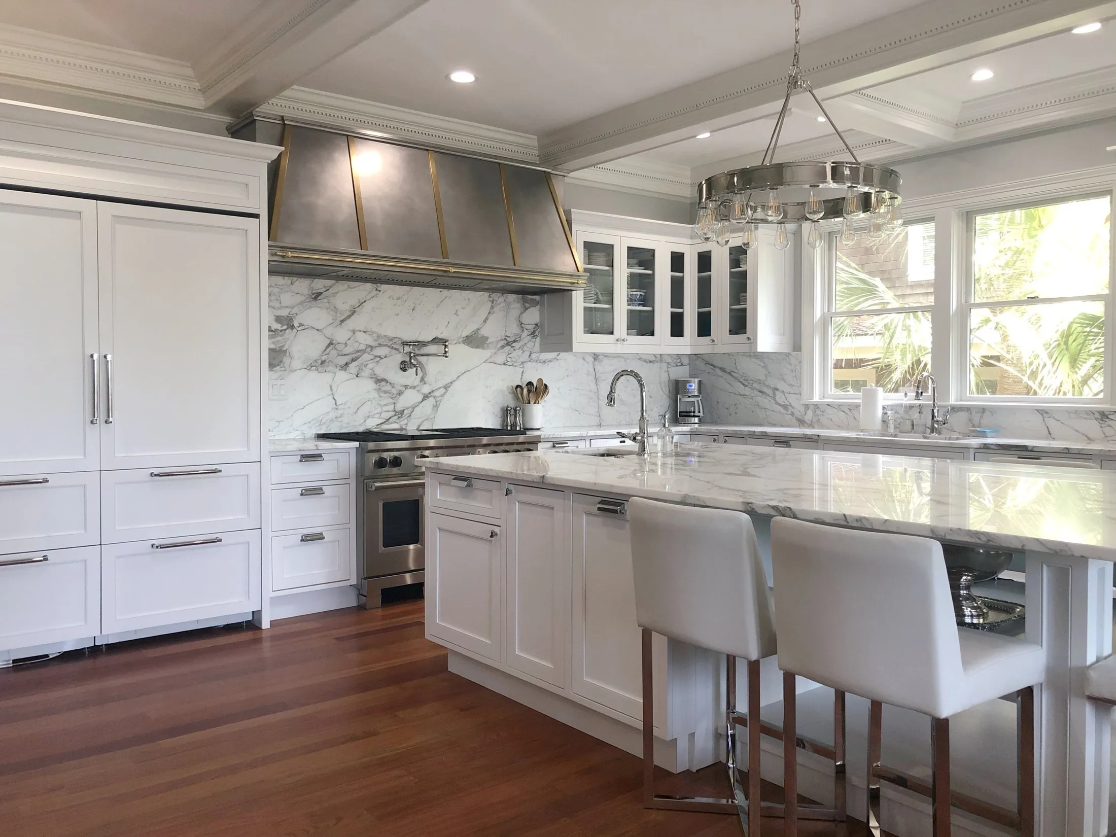 Modern white kitchen with marble countertops, cabinets, and backsplash, stainless steel appliances, a chandelier, and large windows with a view of greenery.