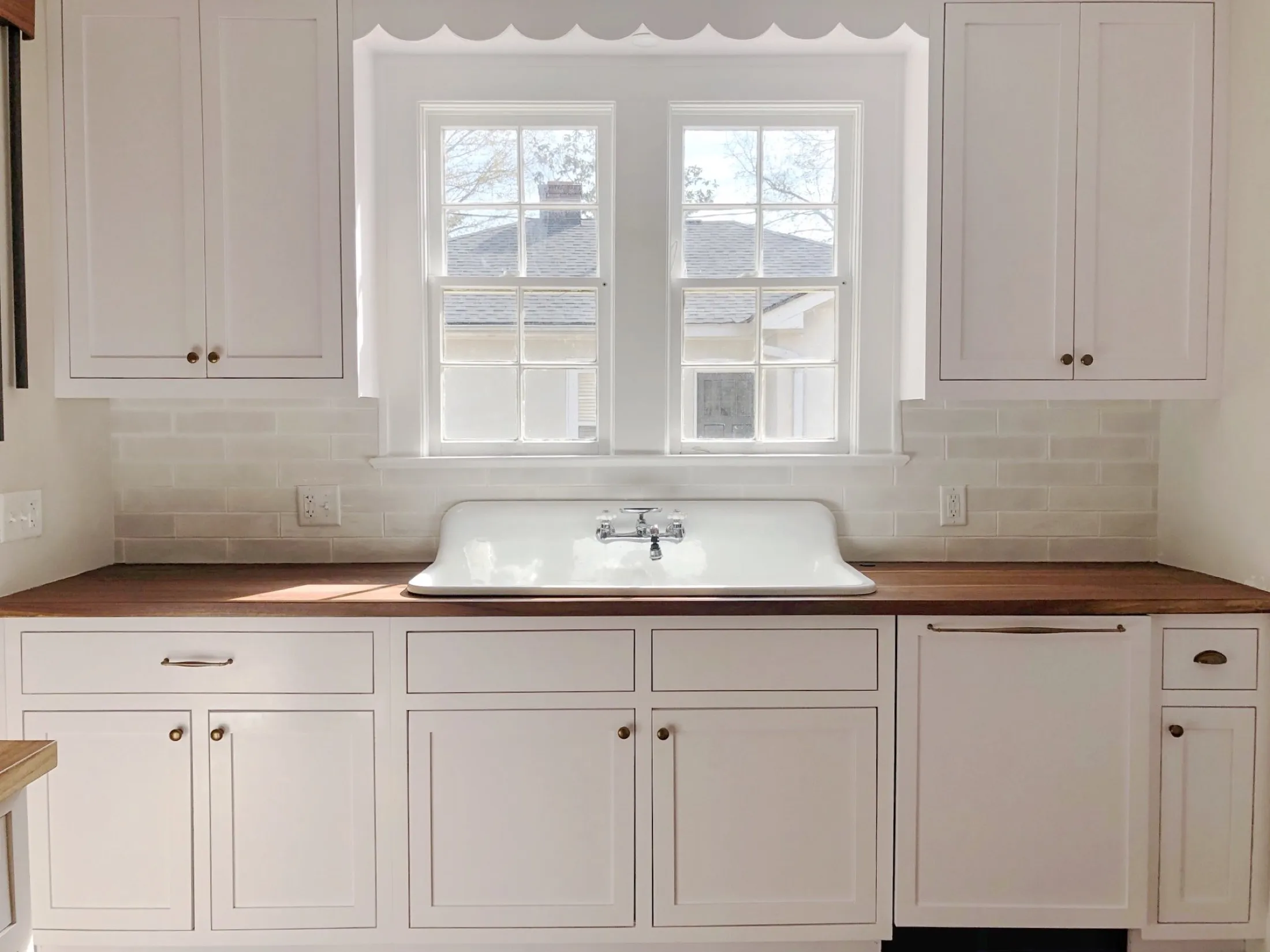 A kitchen with white cabinets, a white backsplash, a wooden countertop, and a white porcelain sink under a large window.