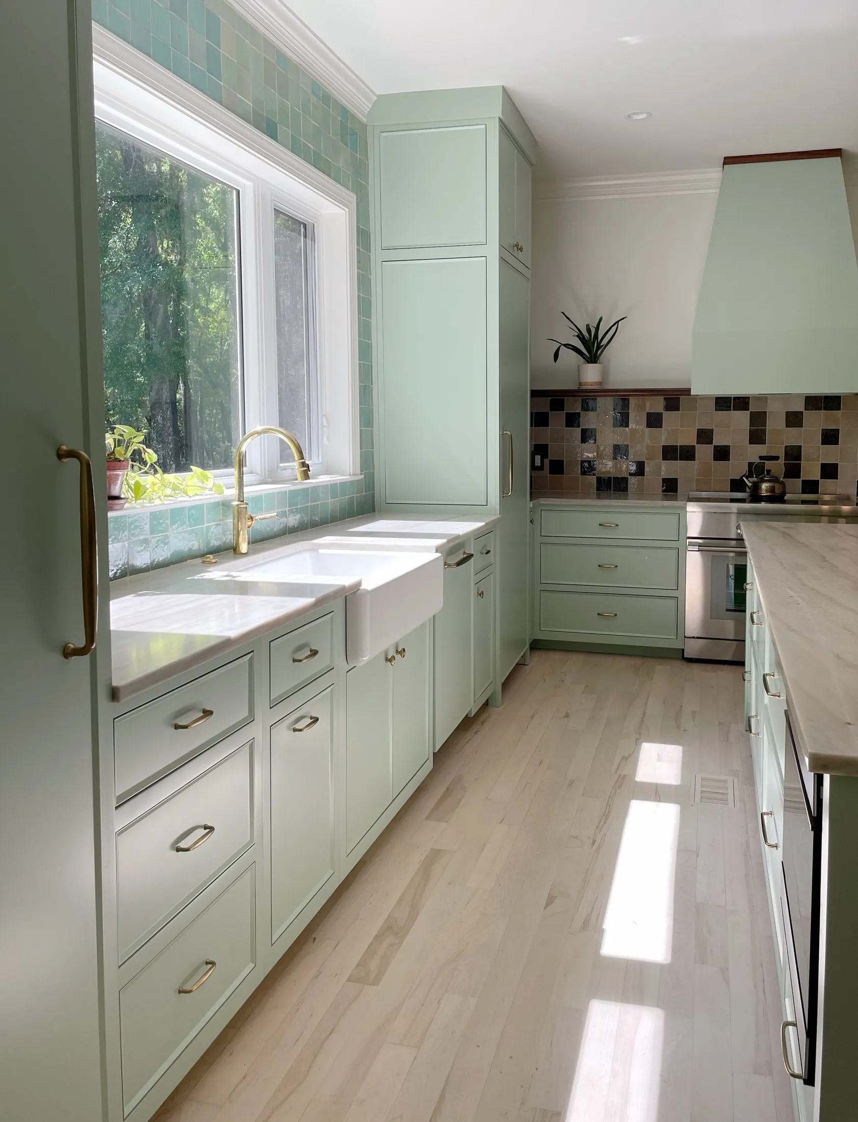 Bright kitchen with mint green cabinets, a farmhouse sink, gold handles, a large window with plants outside, a tiled backsplash, and a stainless steel stove.