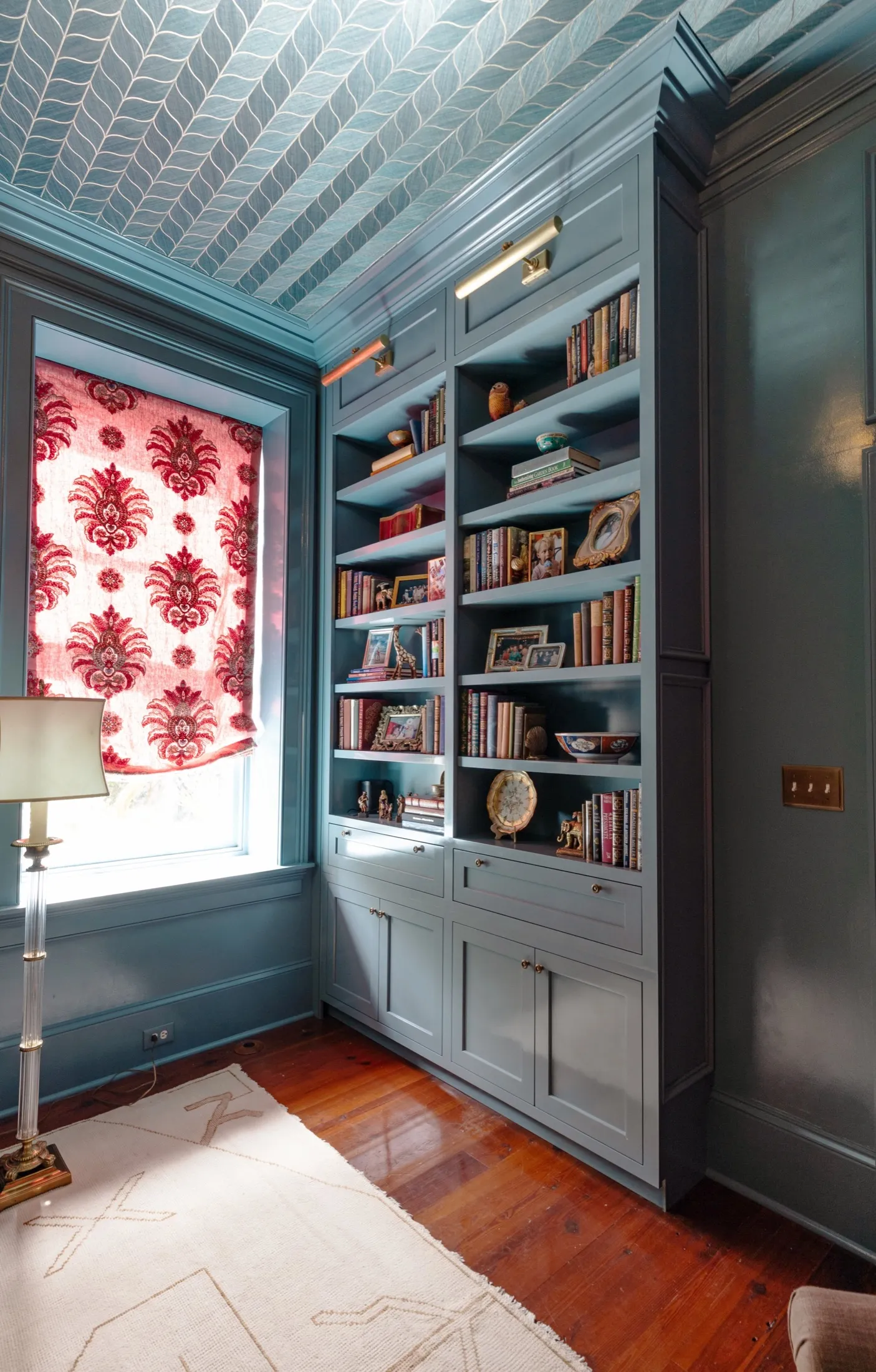 A corner of a room with blue built-in bookshelves filled with books and decorative items, a window with red and white patterned curtains, a beige rug, and a floor lamp, with a wood floor and wall paneling.