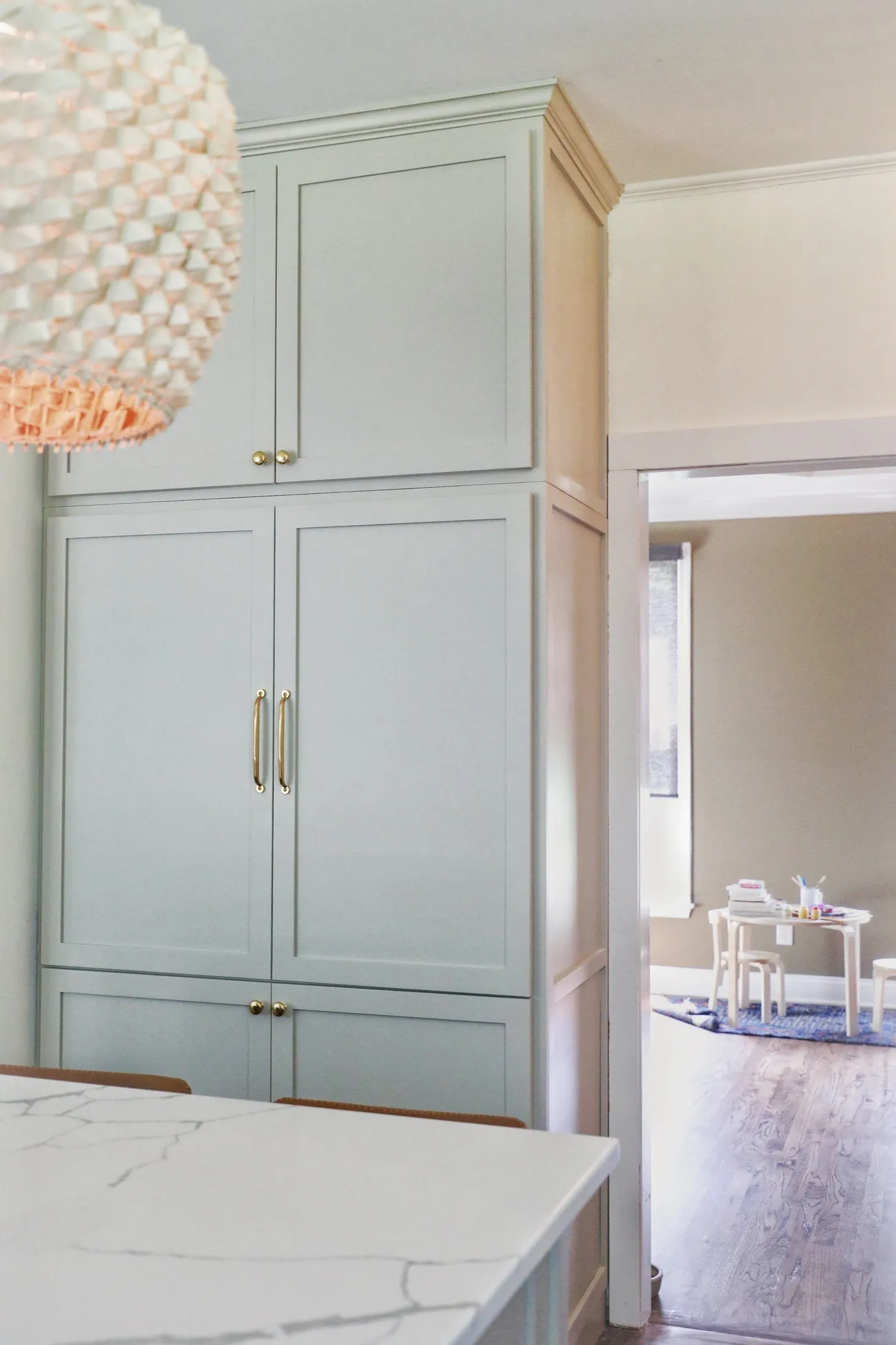 Close-up of a light-colored kitchen cabinet with gold handles, part of a modern kitchen with a marble countertop and a hanging textured light fixture, with a view into a children's playroom with a small table and chairs, stacking cups, and toys.