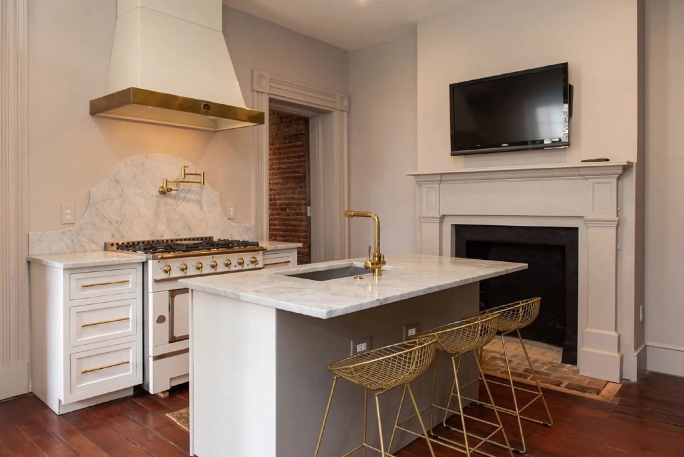 A modern kitchen with white cabinets, a marble countertop and backsplash, a gold kitchen faucet, and gold bar stools. There's a stove with a gold hood above it, a fireplace with a TV mounted above, and hardwood floors.