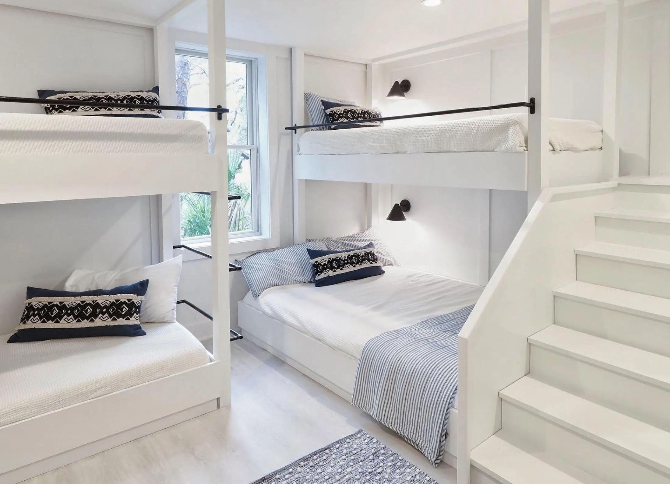 A bright bedroom with white walls and furniture, featuring a double bed with striped bedding and pillows, and two sets of bunk beds with black railings and black-patterned pillows, a window with a view of greenery.