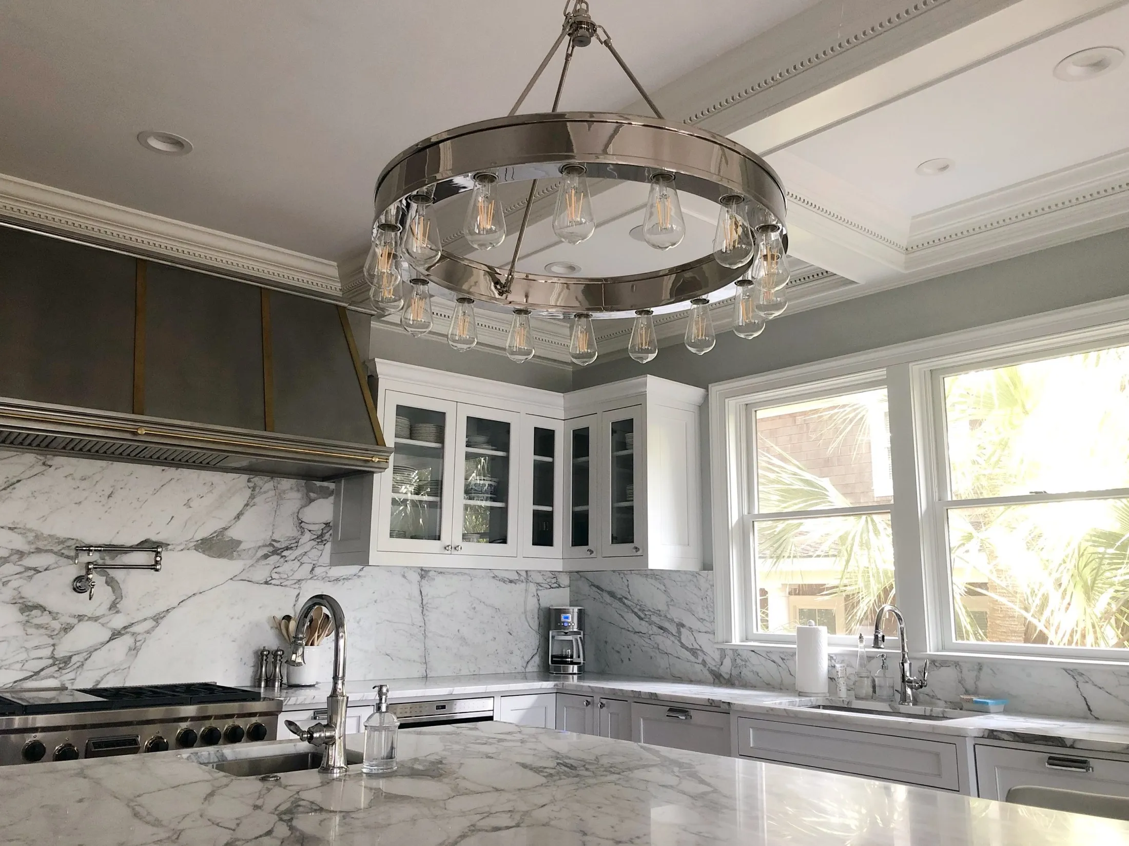 Modern kitchen with marble countertops and backsplash, white cabinets, large windows, and a circular chandelier with exposed bulbs.