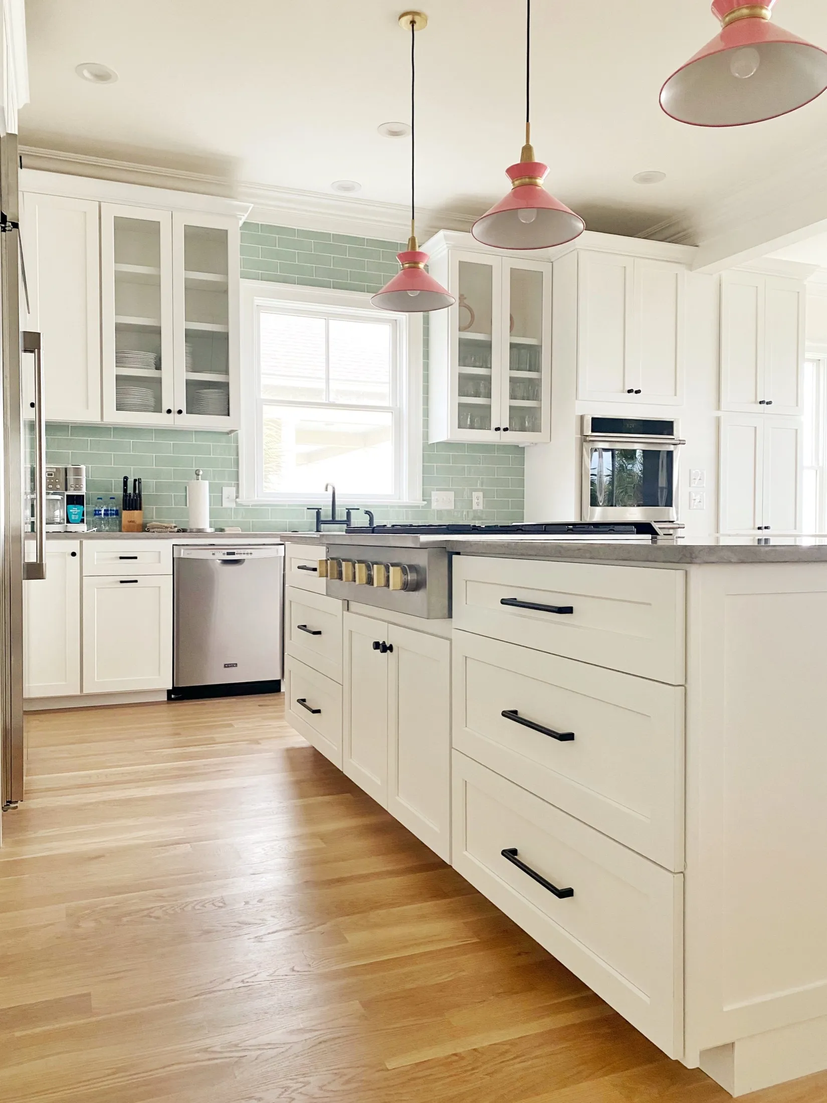 Bright, modern kitchen with white cabinets, light green subway tile backsplash, wooden floors, and pink pendant lights.