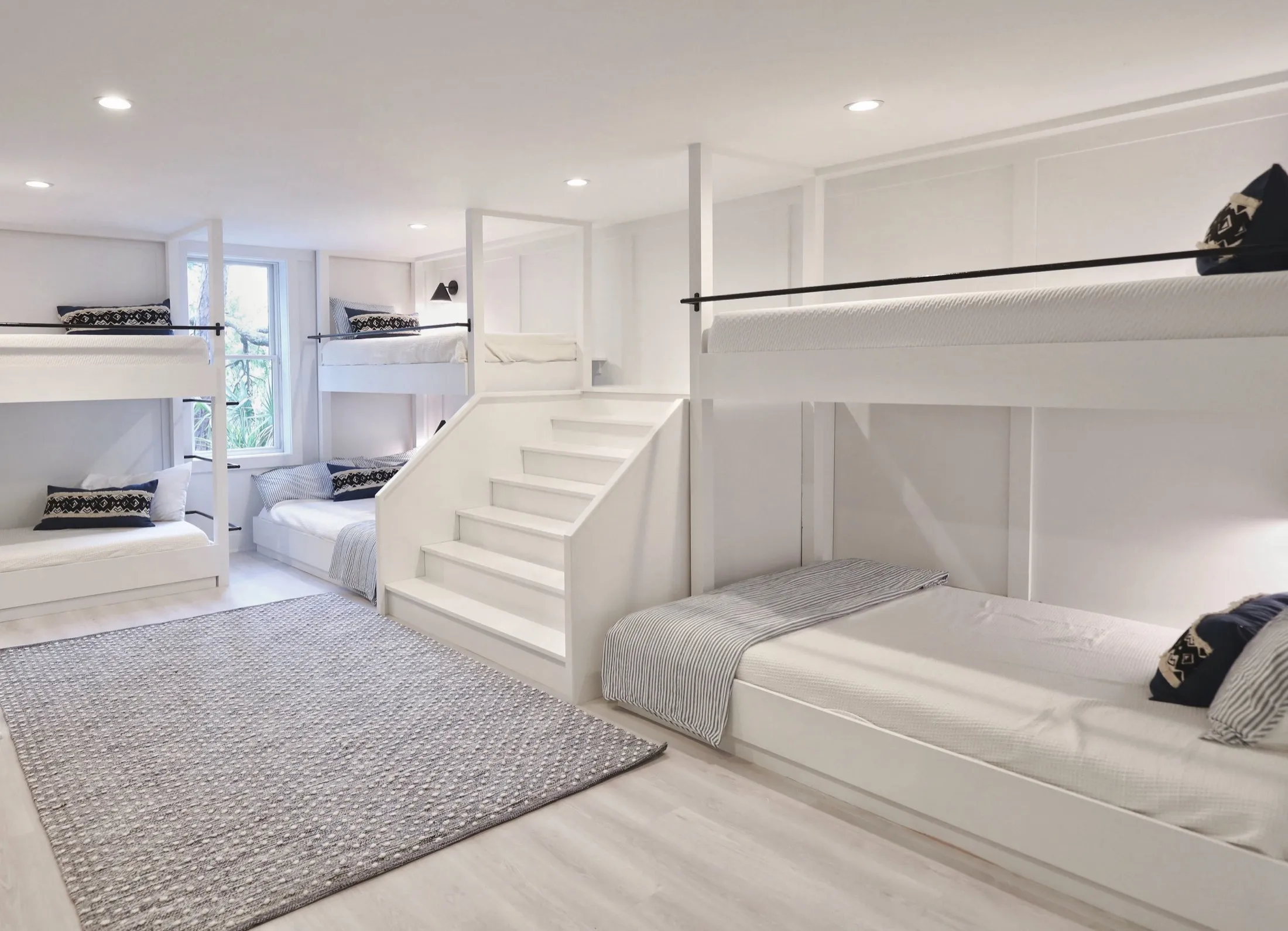 Interior of a modern bedrooms with white bunk beds, black and white pillows, a gray area rug, and small windows, illuminated by recessed ceiling lights.