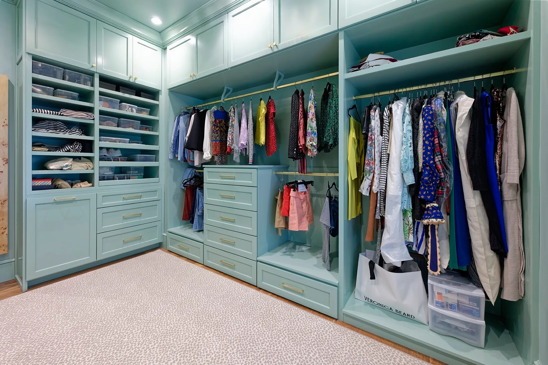 A walk-in closet with mint green cabinetry and shelves filled with folded clothes, shoe boxes, and hanging clothes, under bright lighting.