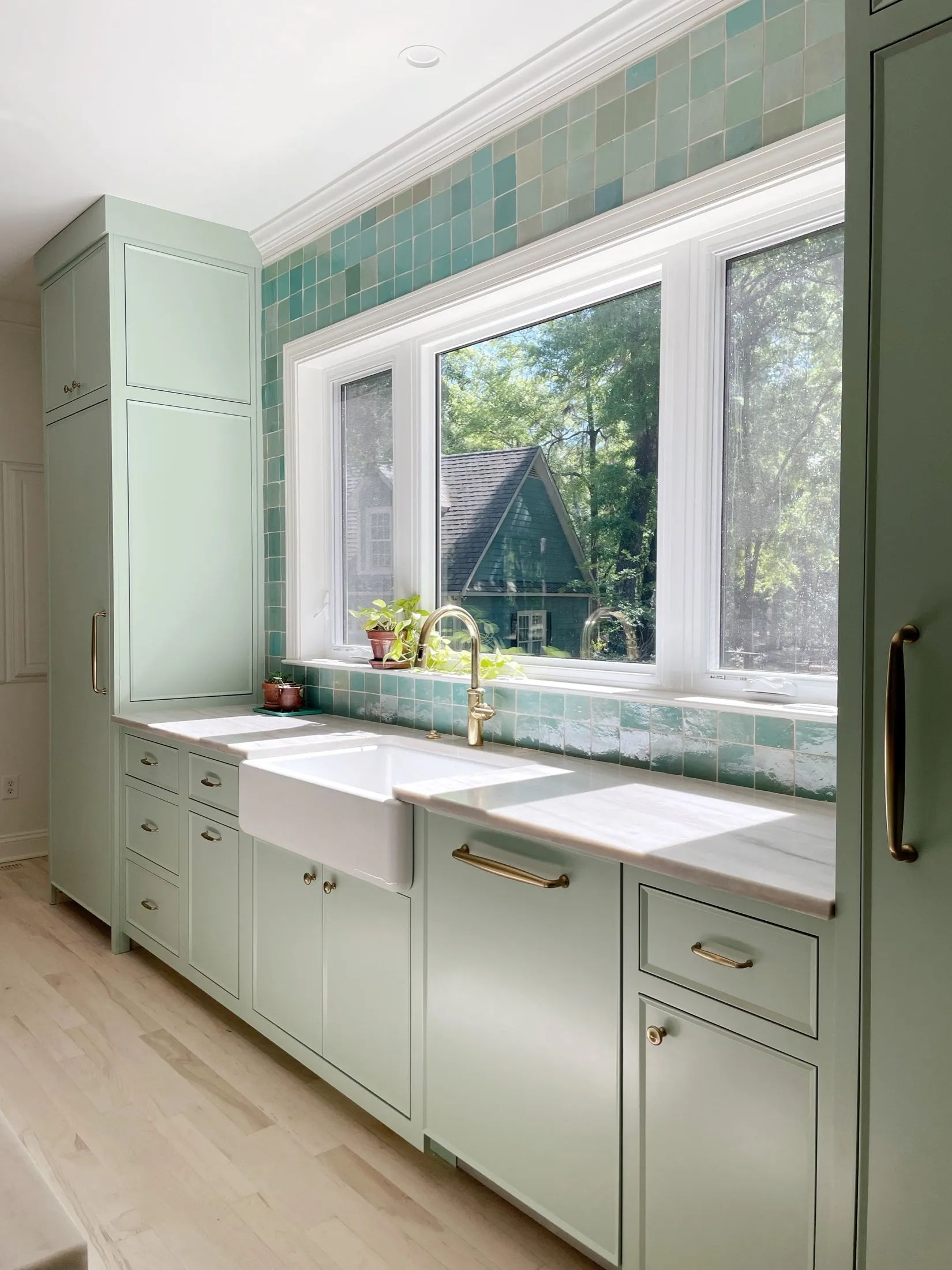 A bright kitchen with light green cabinets, a double window above a white farmhouse sink, and a backsplash of green tiles. Outside the window, trees and a neighboring house are visible.