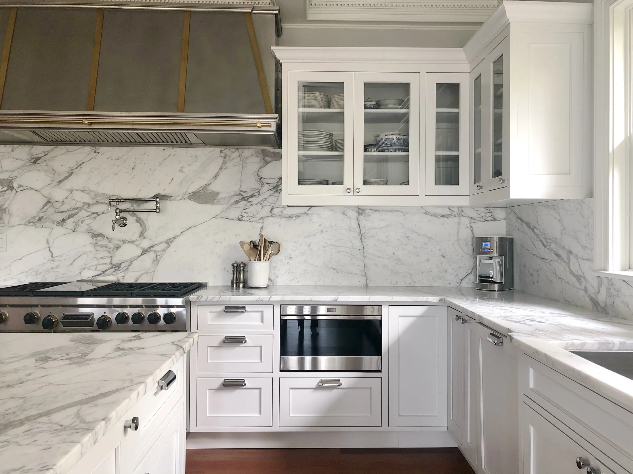 A modern kitchen with white cabinetry, marble countertops and backsplash, a stainless steel oven, a coffee maker near the window, and a dishware cabinet with glass doors.