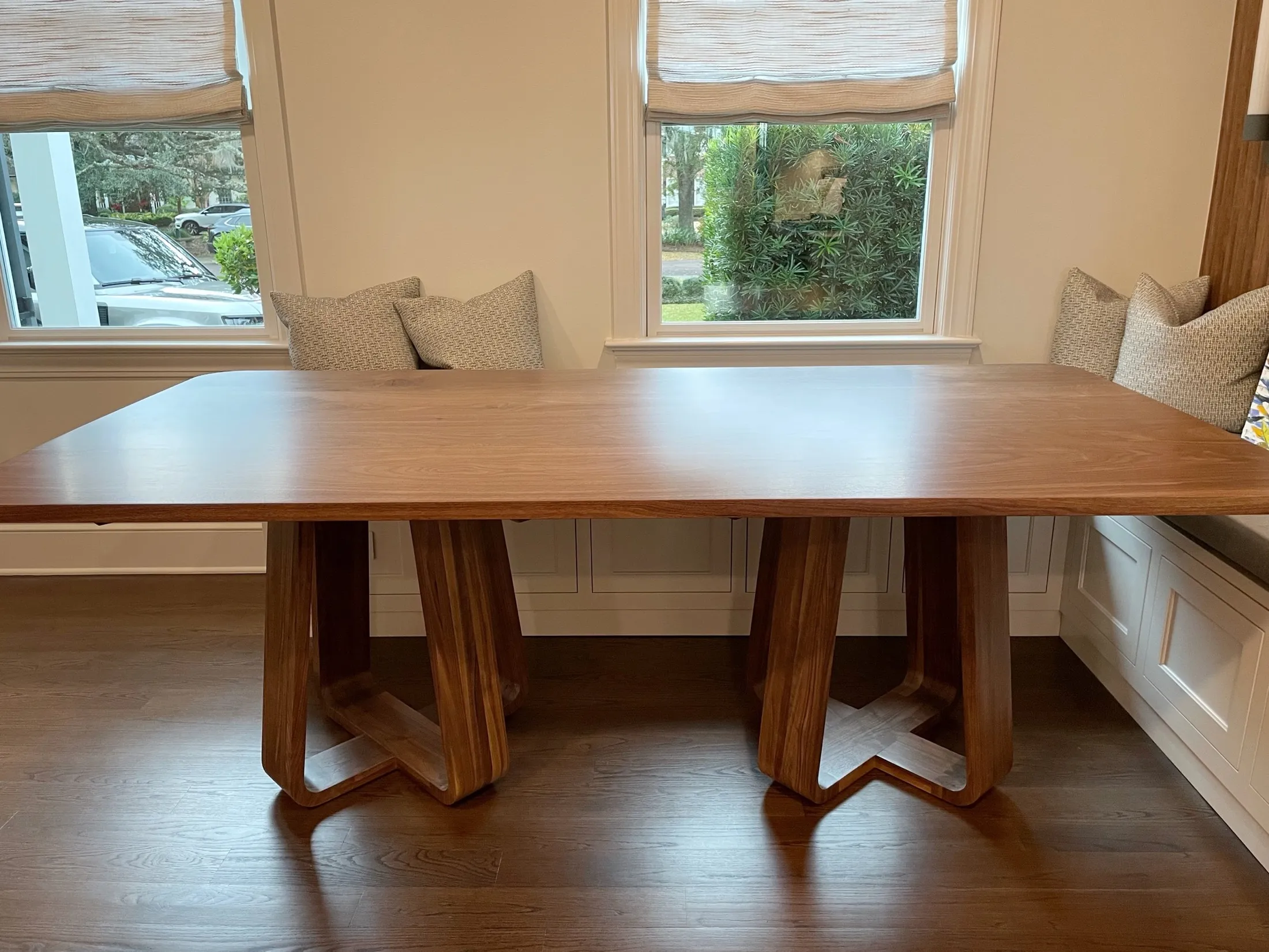 A wooden dining table with modern, thick legs in a room with hardwood floors, two windows with beige blinds, and beige and patterned pillows on the window seats.