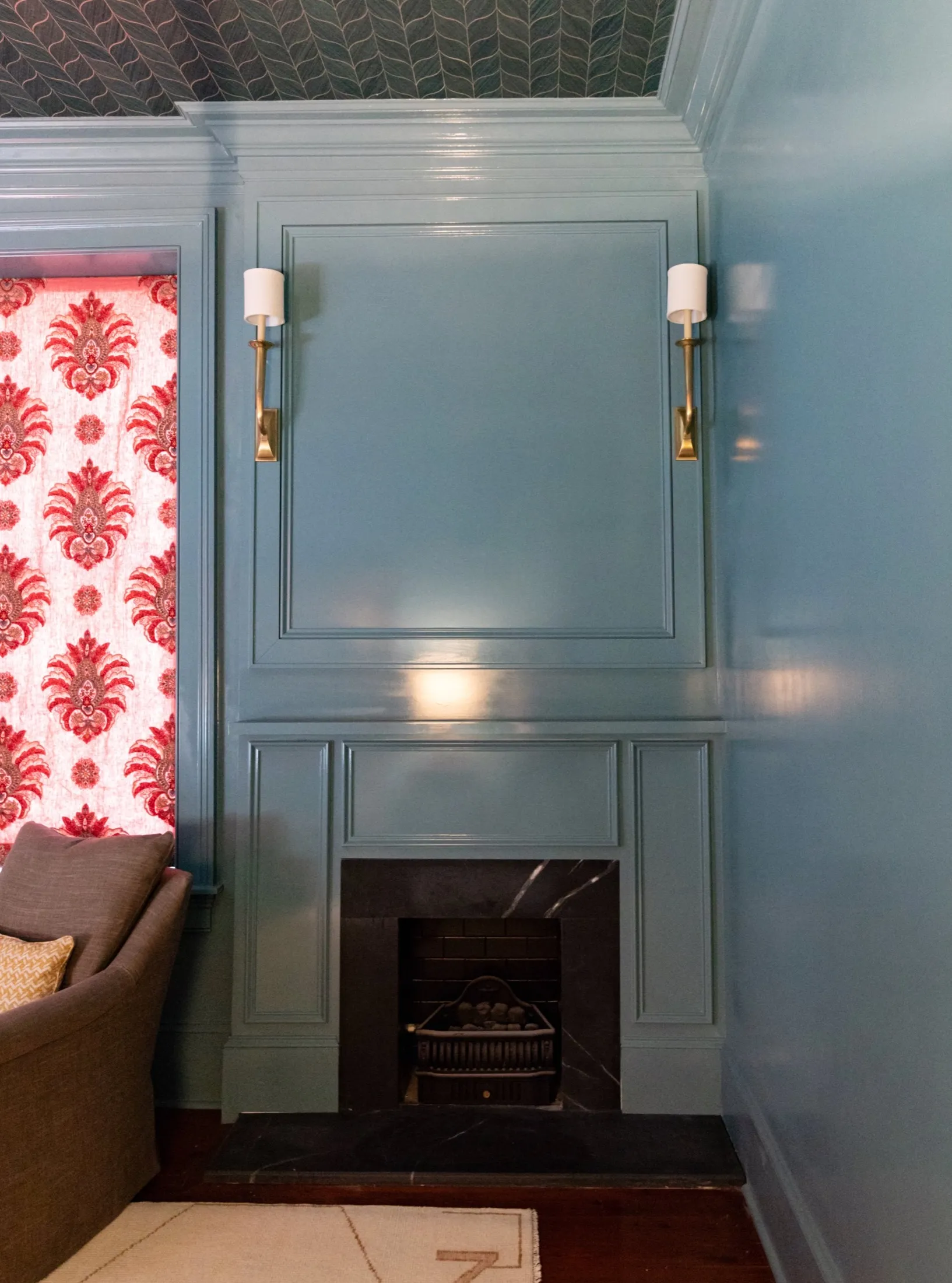 Interior living room with blue paneled fireplace wall, brass wall sconces, and a brown sofa with a yellow pillow. Red patterned curtain is partially visible on the left.