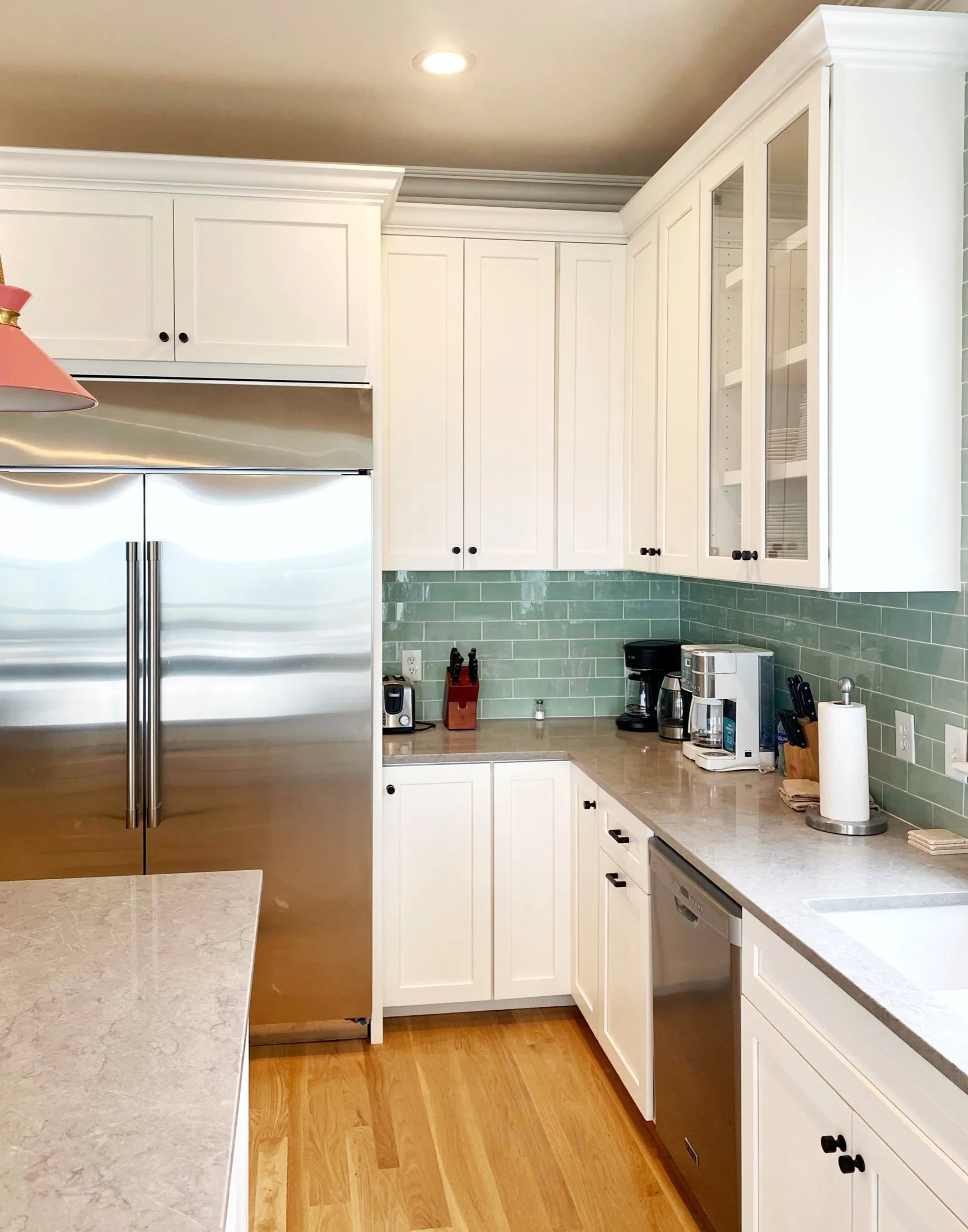 Kitchen with white cabinets, green tile backsplash, stainless steel refrigerator, countertop with coffee maker, toaster, paper towels, and knives, hardwood floor.