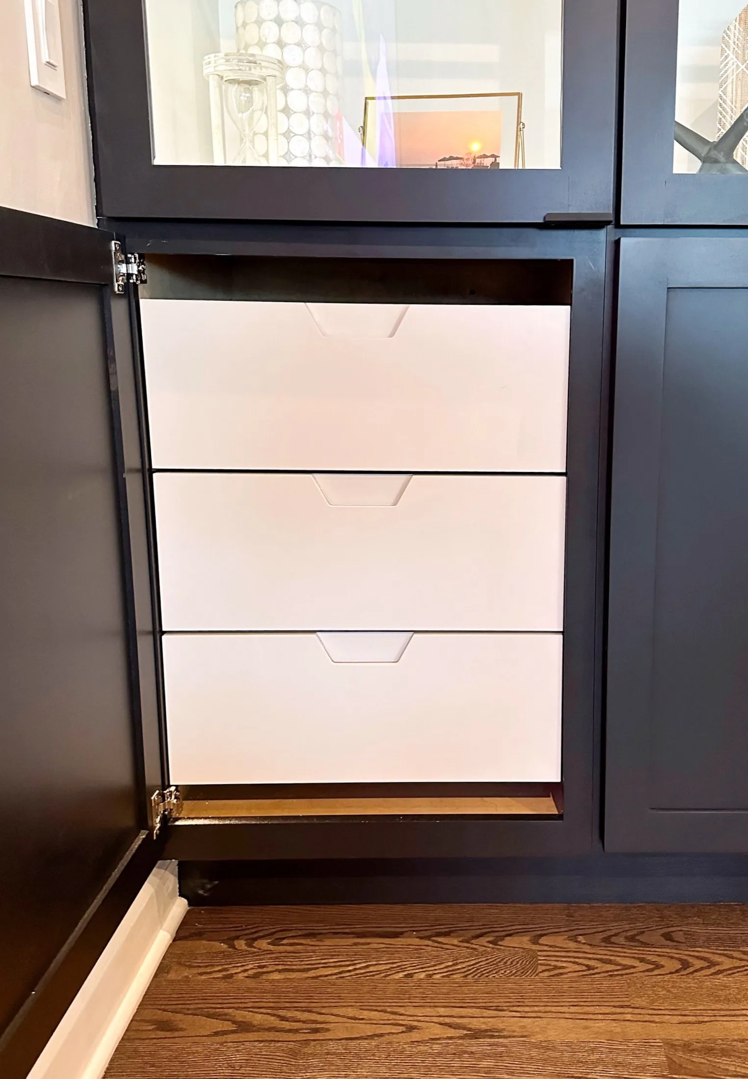View of a kitchen cabinet with three white drawers beneath a black countertop, on a wooden floor.