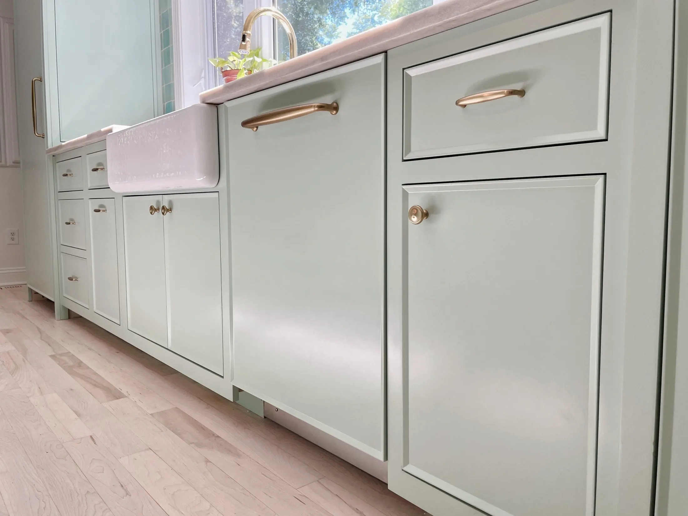Light green kitchen cabinets with rose gold handles, light-colored wood floor, and a farmhouse sink with a window above.