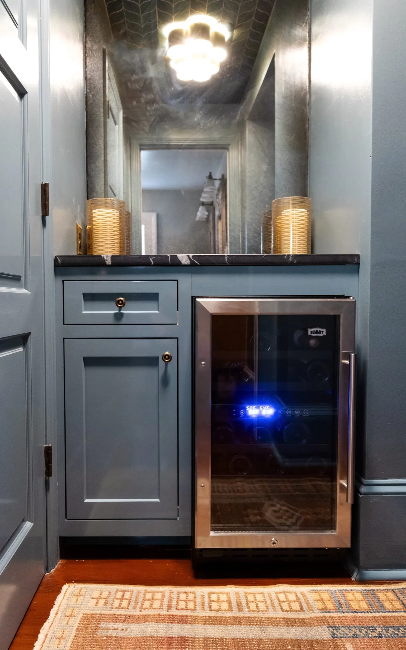 A built-in wine cooler with a glass door, gray cabinetry with brass knobs, a marble countertop, and decorative lamps, reflected in a large mirror on the wall.
