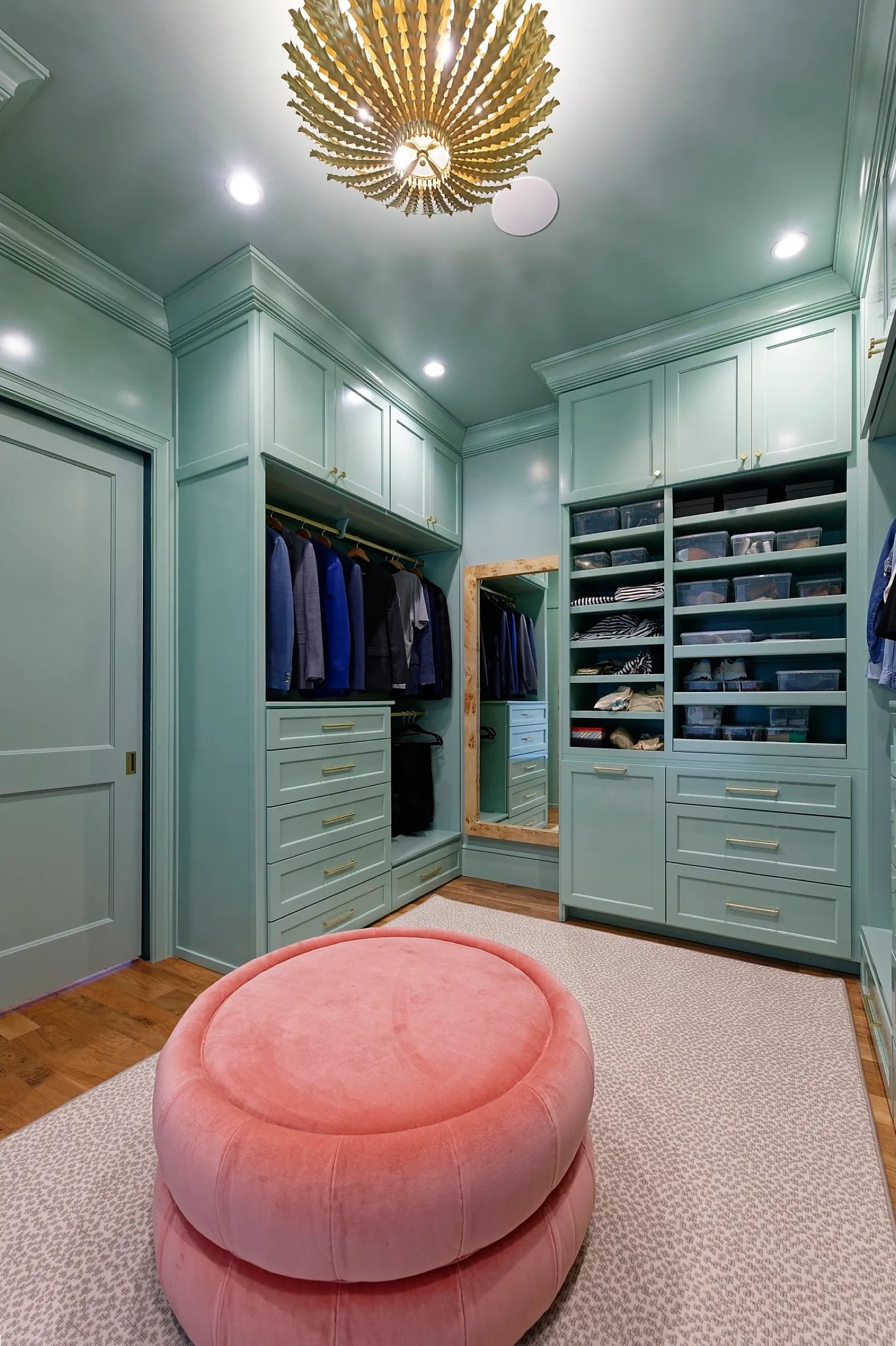 Walk-in closet with mint green built-in cabinets, a pink velvet ottoman, a large mirror, and clothing organized in drawers and shelves, with a decorative gold chandelier and ceiling lights.