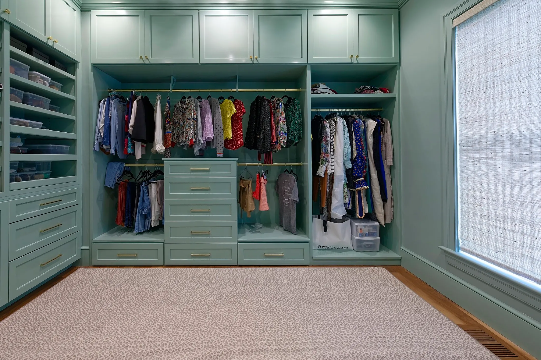 Walk-in closet with mint green cabinetry, clothing hanging on gold rods, organized drawers, and a window with woven shade, beige carpet