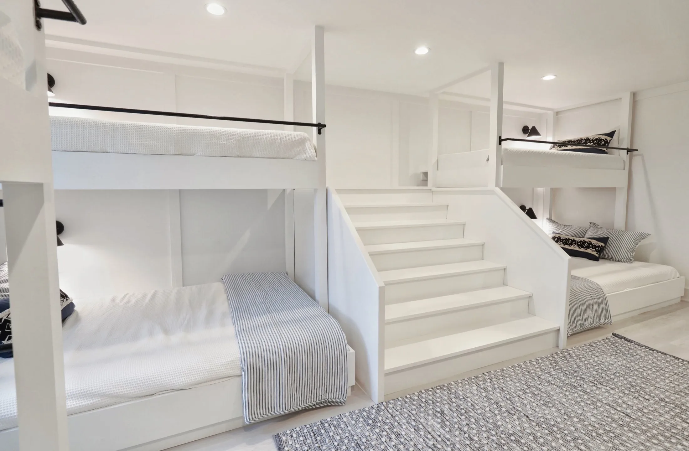 A white staircase with three steps leading to bunk beds on either side, each with black and white bedding and pillows, in a minimalistic, modern bedroom with white walls and ceiling, recessed lighting, and a textured area rug on the floor.