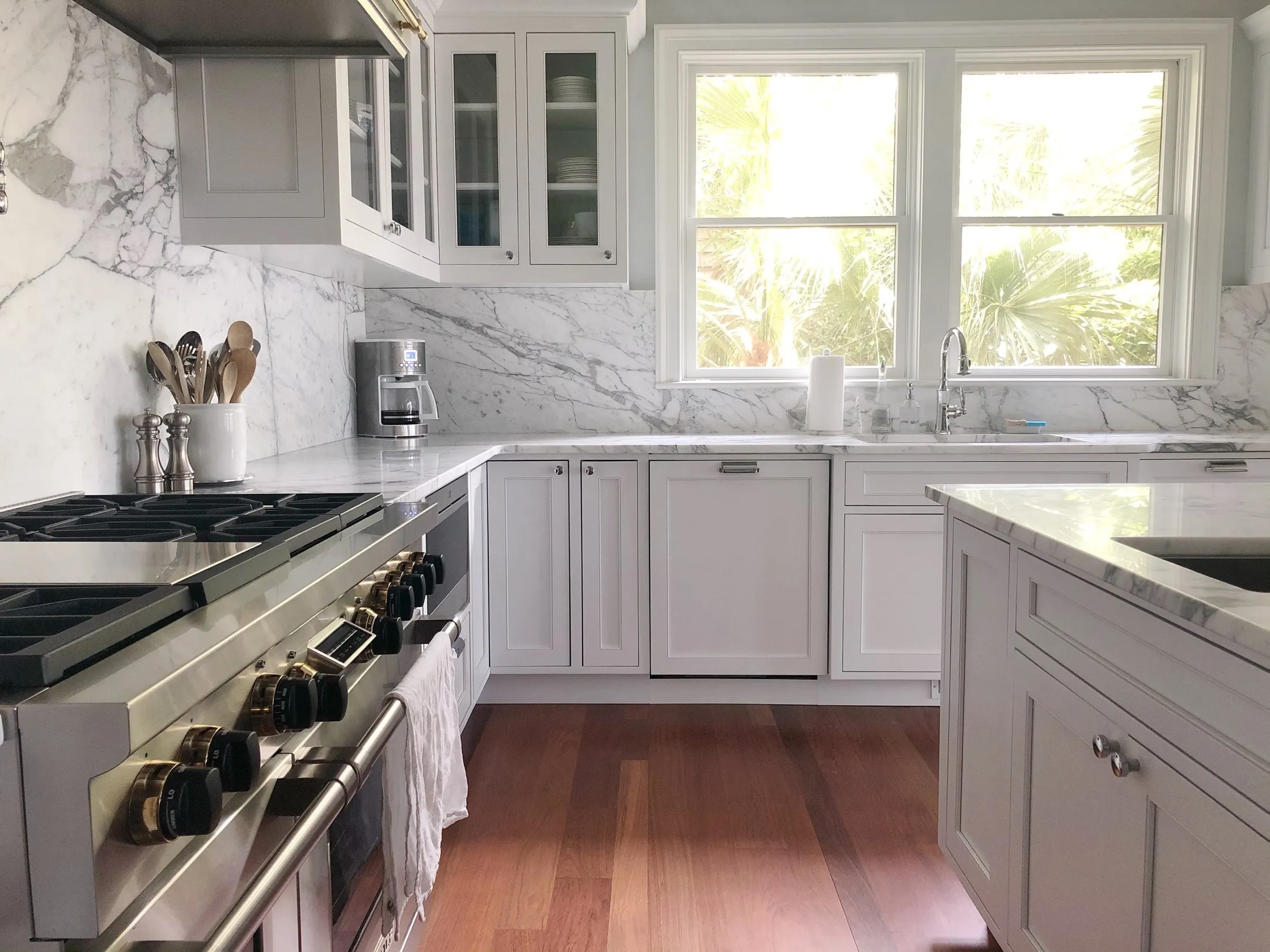Bright kitchen with white cabinets, marble countertops, and backsplash, stainless steel stove, coffee maker, paper towel roll, and large windows showing palm trees outside.