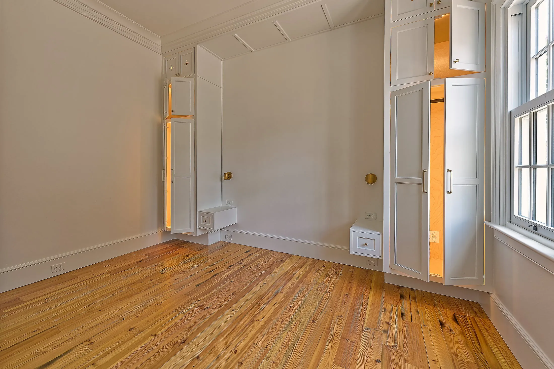 Built-in armoire with its door open to a lit interior beside a tall window and heart-pine floors.