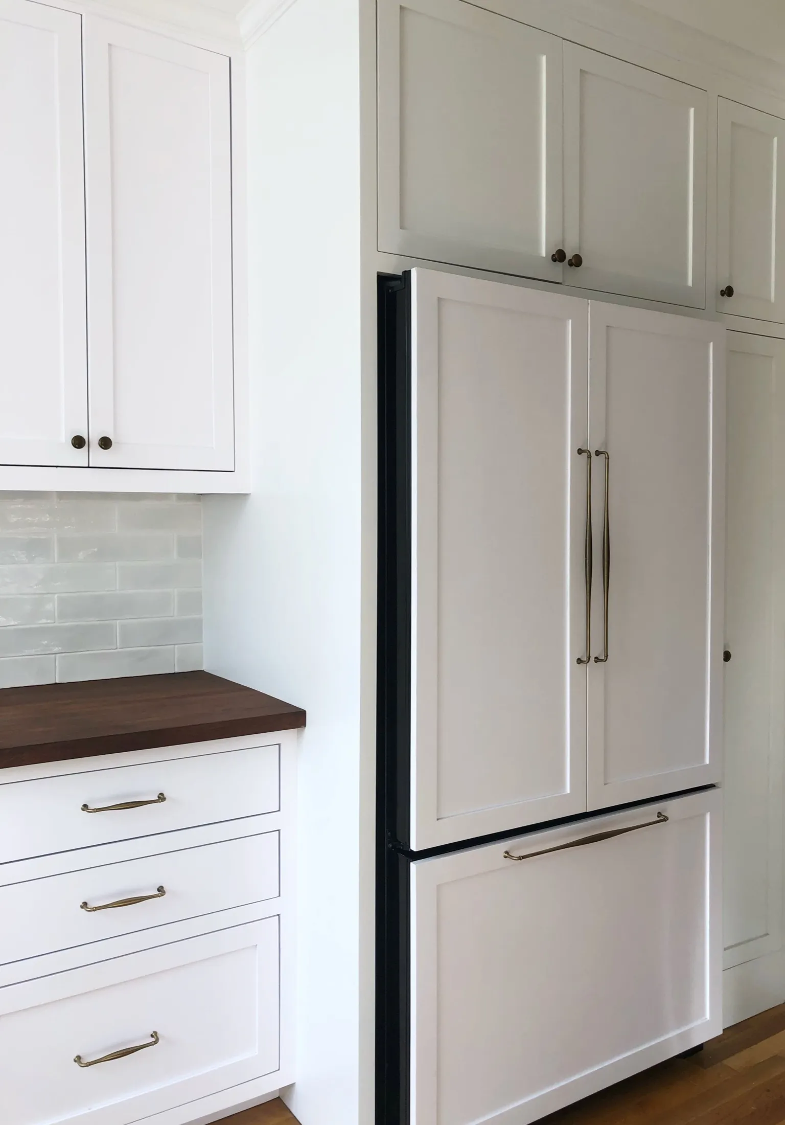 White kitchen cabinets, a dark countertop, and a large white refrigerator with brass handles against a white subway tile backsplash.