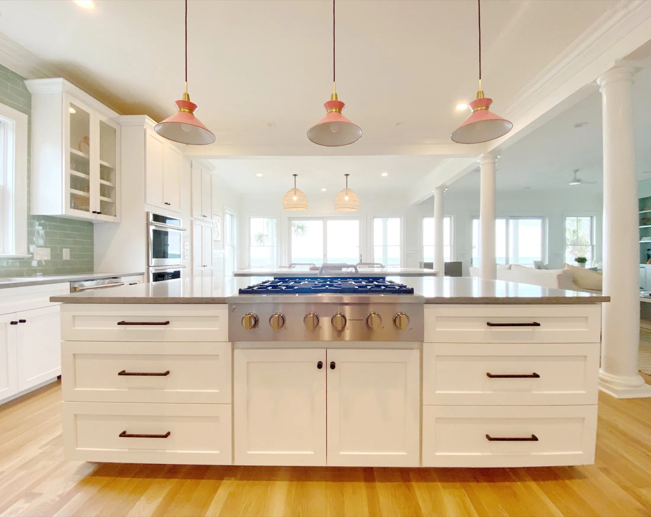 Bright kitchen with white cabinets, an island with a stove, wooden flooring, and pendant lights, overlooking a living area with large windows and ocean view.