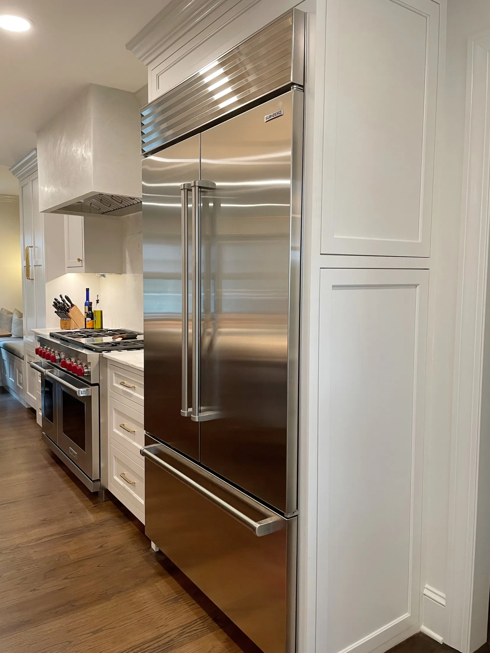 Stainless steel refrigerator in a modern kitchen with white cabinets, a gas stove, and wooden flooring.